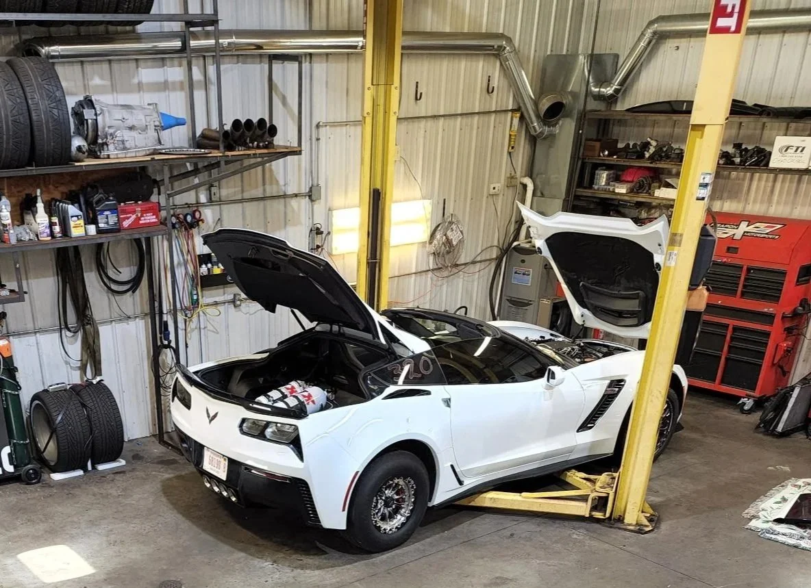 White sports car in a garage with hood and trunk open, car lift underneath, and tools and shelves on the walls.