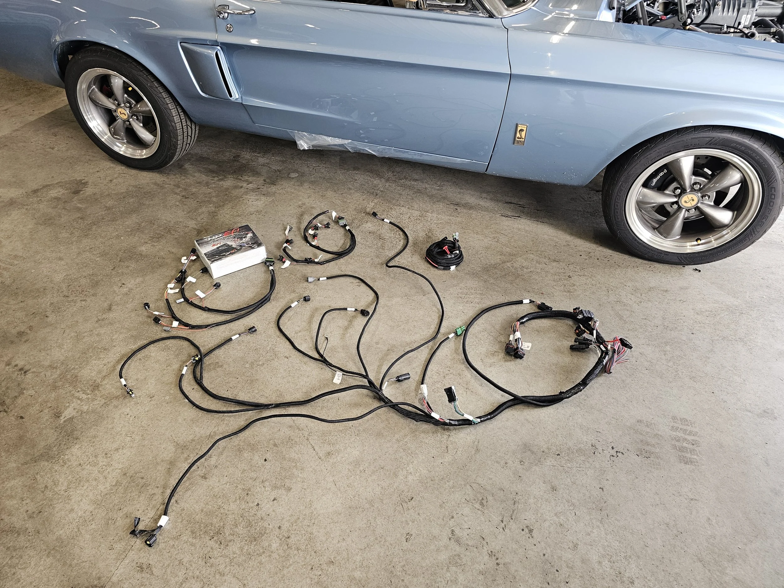 A vintage blue Ford Shelby Mustang car parked on a concrete floor with an engine wiring harness spread out on the ground in front of it.