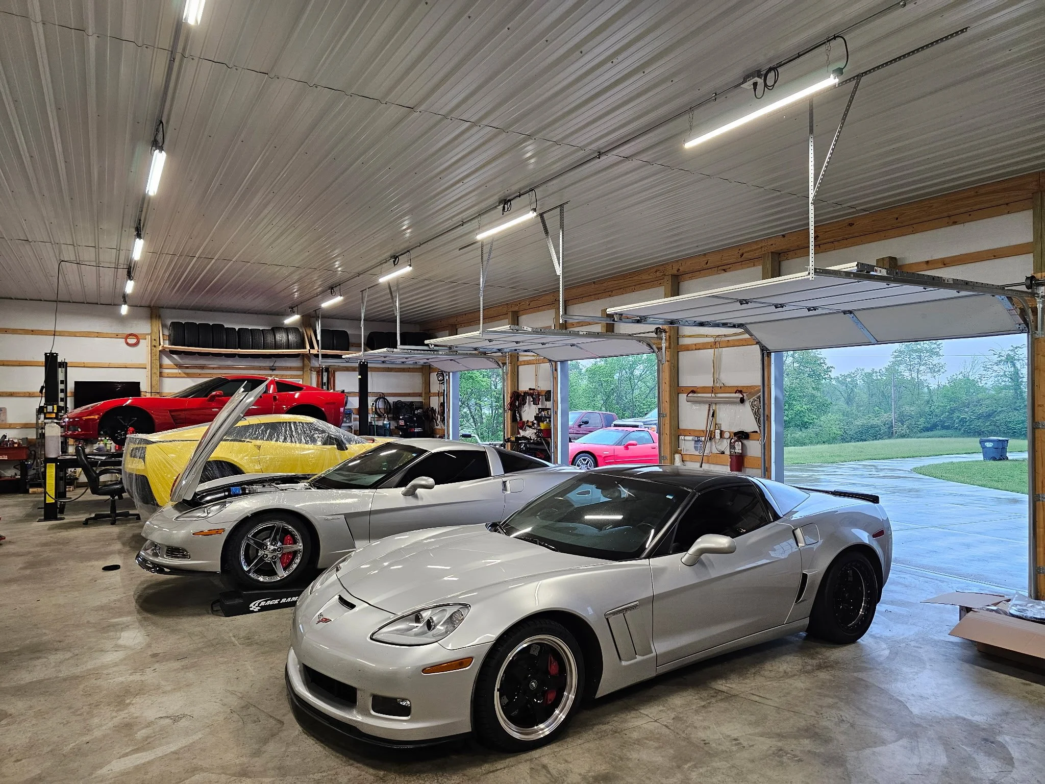 A garage with four sports cars, including a silver Corvette in the foreground and a red Lamborghini on a lift, with another yellow car and red car in the background and open garage doors showing the outdoors.