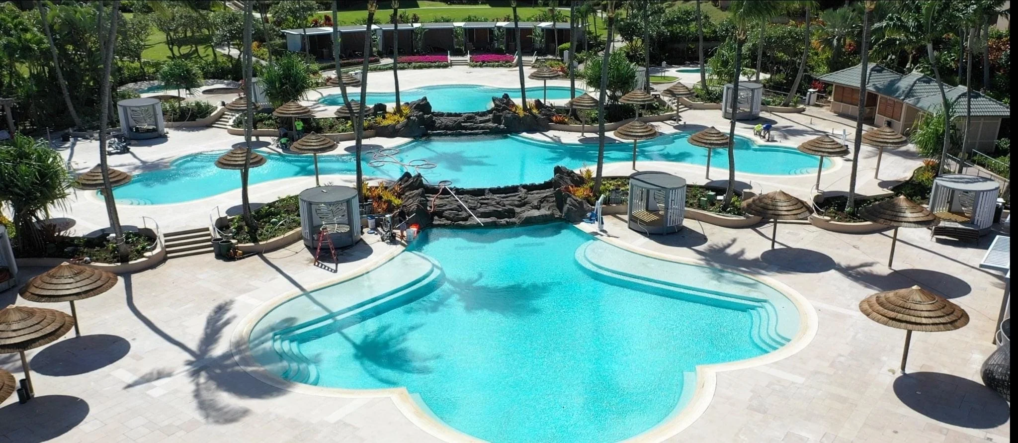 An aerial view of a resort pool area with multiple interconnected swimming pools, tropical trees, and numerous bamboo umbrellas providing shade