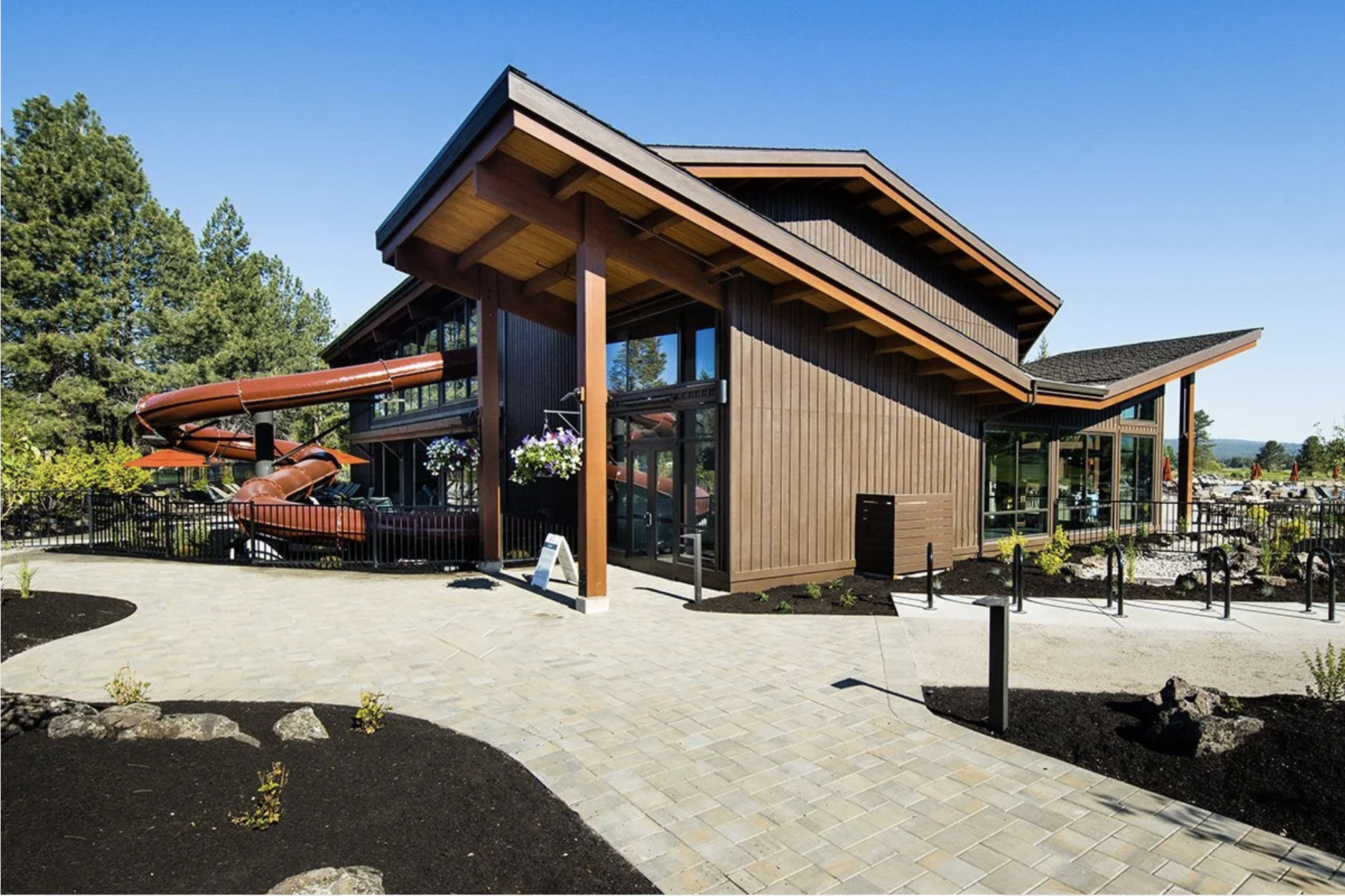 A modern wooden building with large glass windows and multiple sloped roofs, surrounded by trees and a paved patio with outdoor seating.