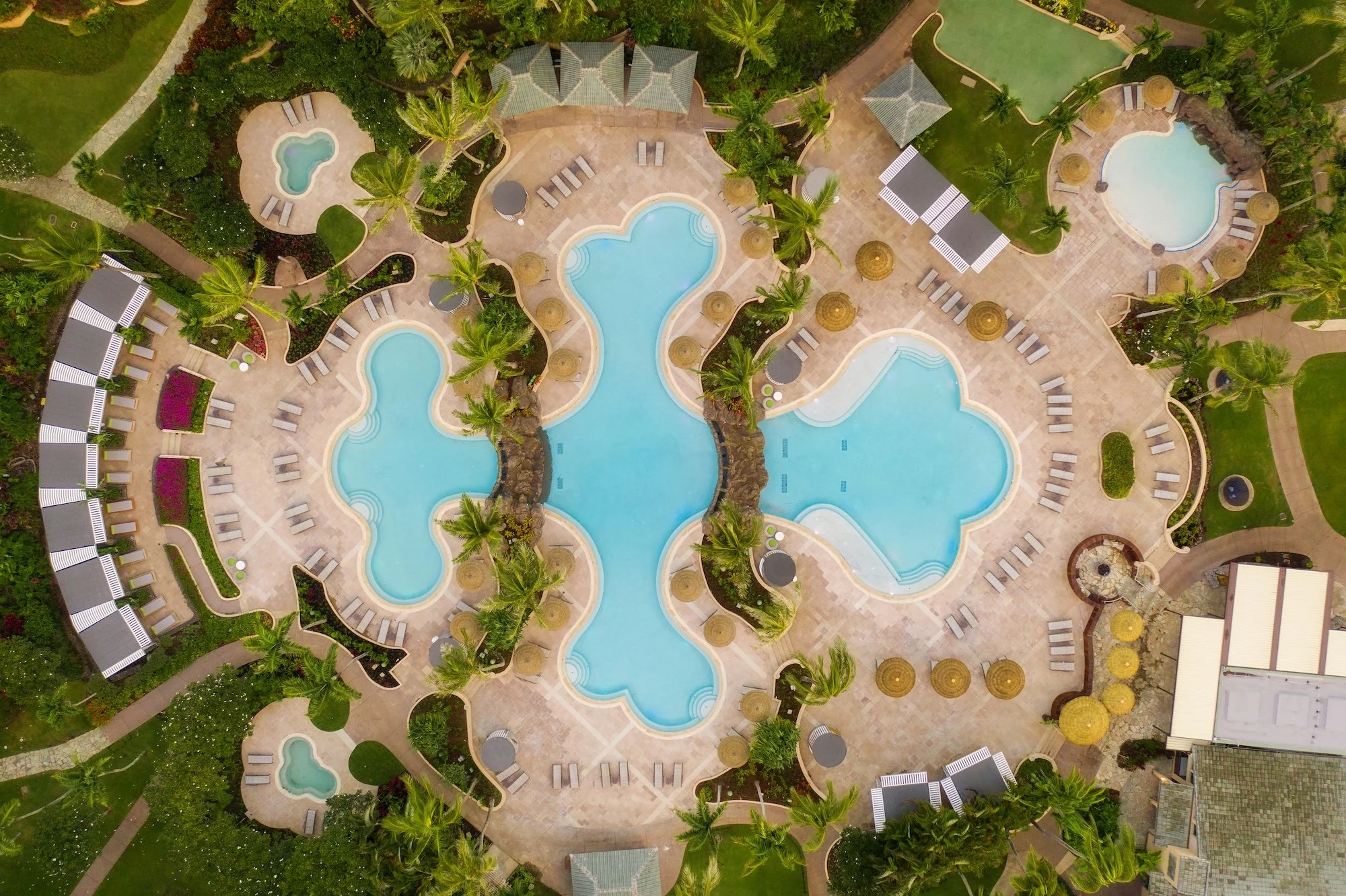 An aerial view of a resort pool area with multiple pools, surrounded by palm trees and lounge chairs.