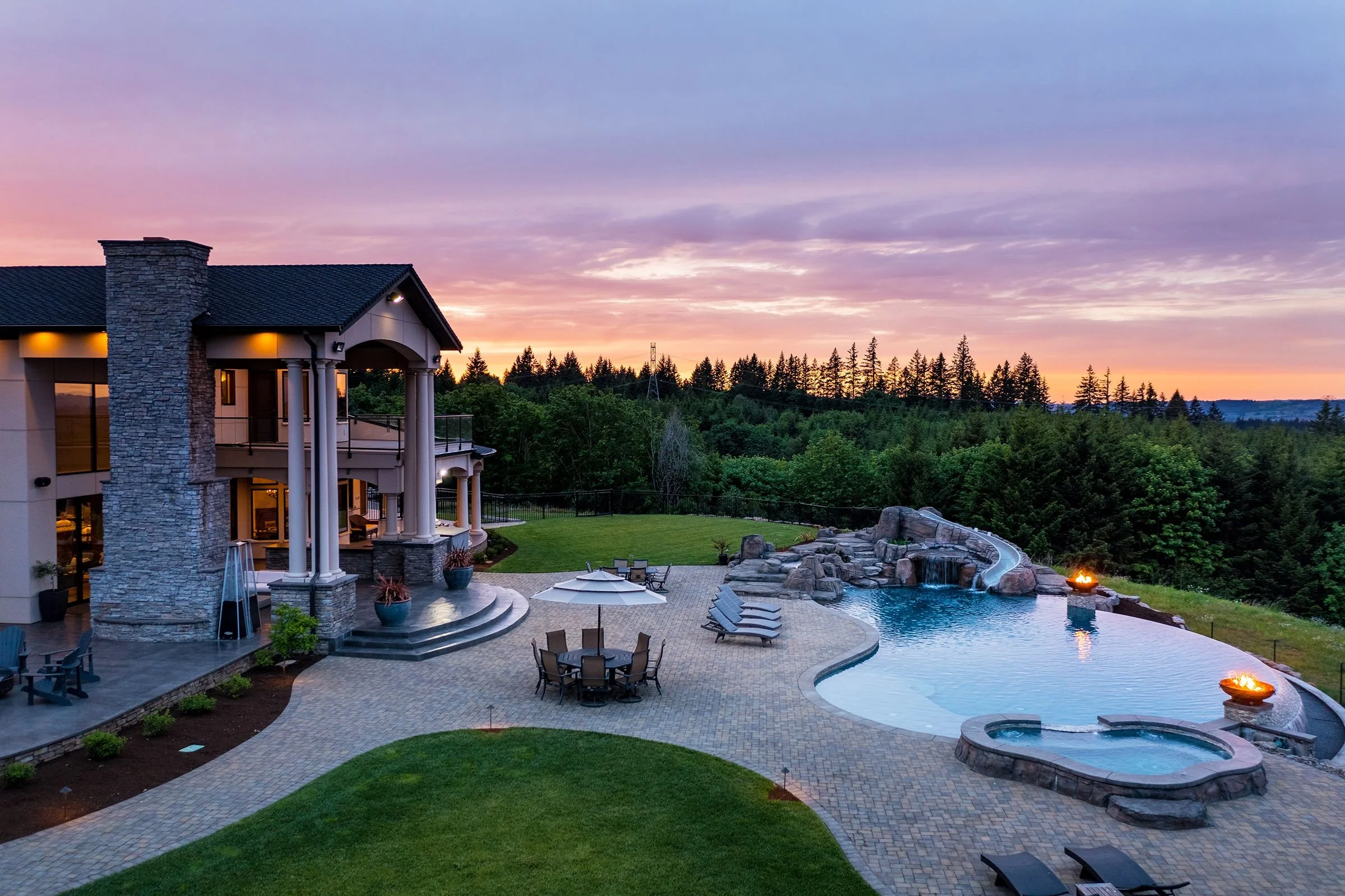 A luxurious house with an outdoor pool and hot tub at sunset, surrounded by a lush green landscape and trees.