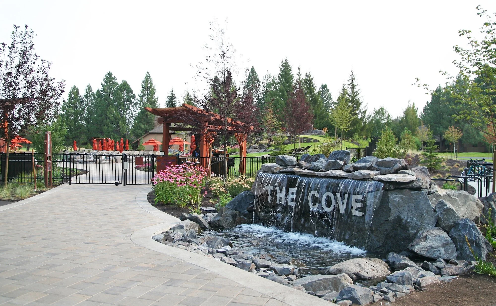 A park with a water feature, a waterfall flowing over rocks with the word "Welcome" on a large stone. There are trees, a walking path, and outdoor seating with umbrellas in the background.