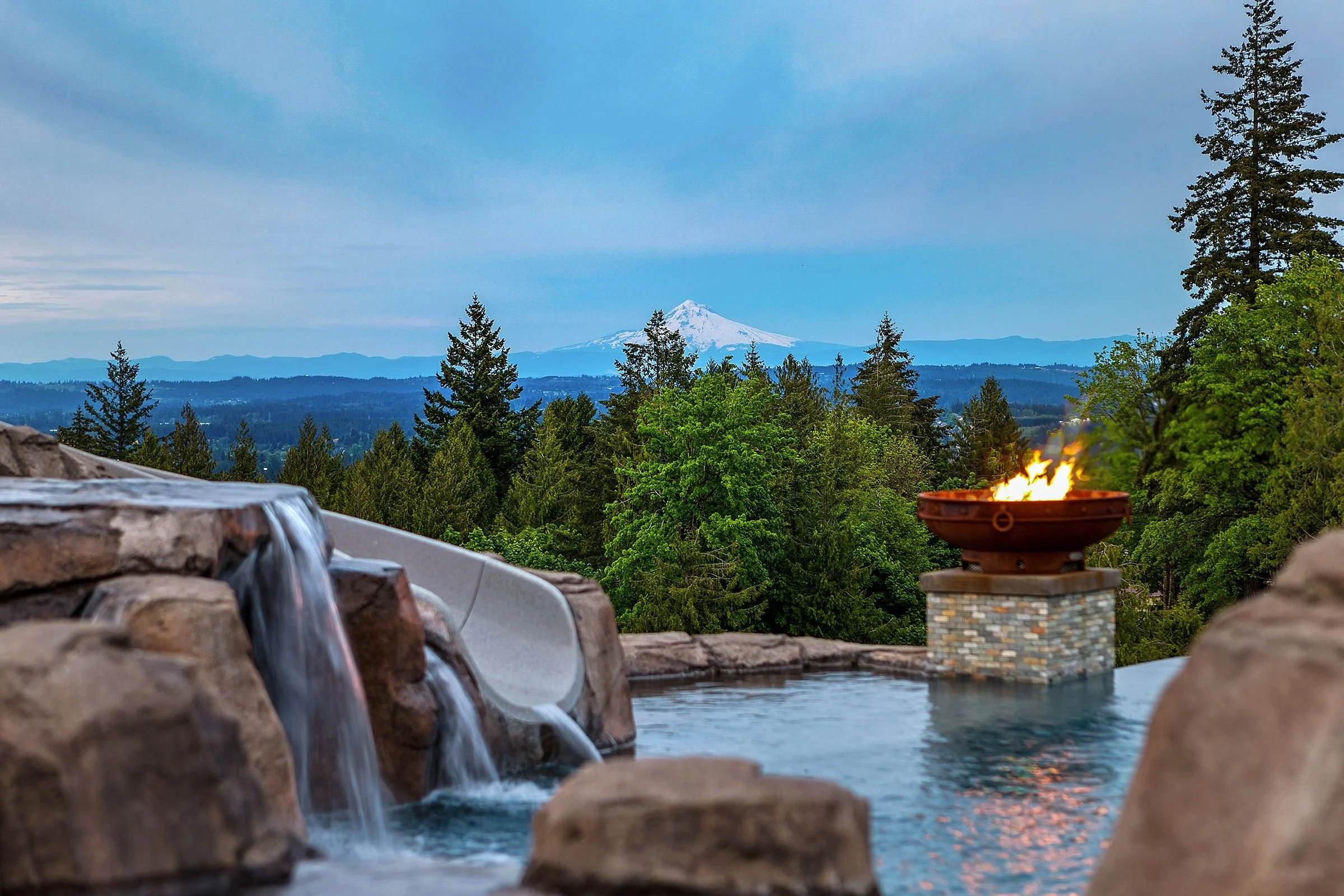 A scenic outdoor view featuring a mountain in the background, surrounded by a forest of tall green trees. In the foreground, there is a landscaped area with a water feature, a fire pit with flames, and natural stone accents.