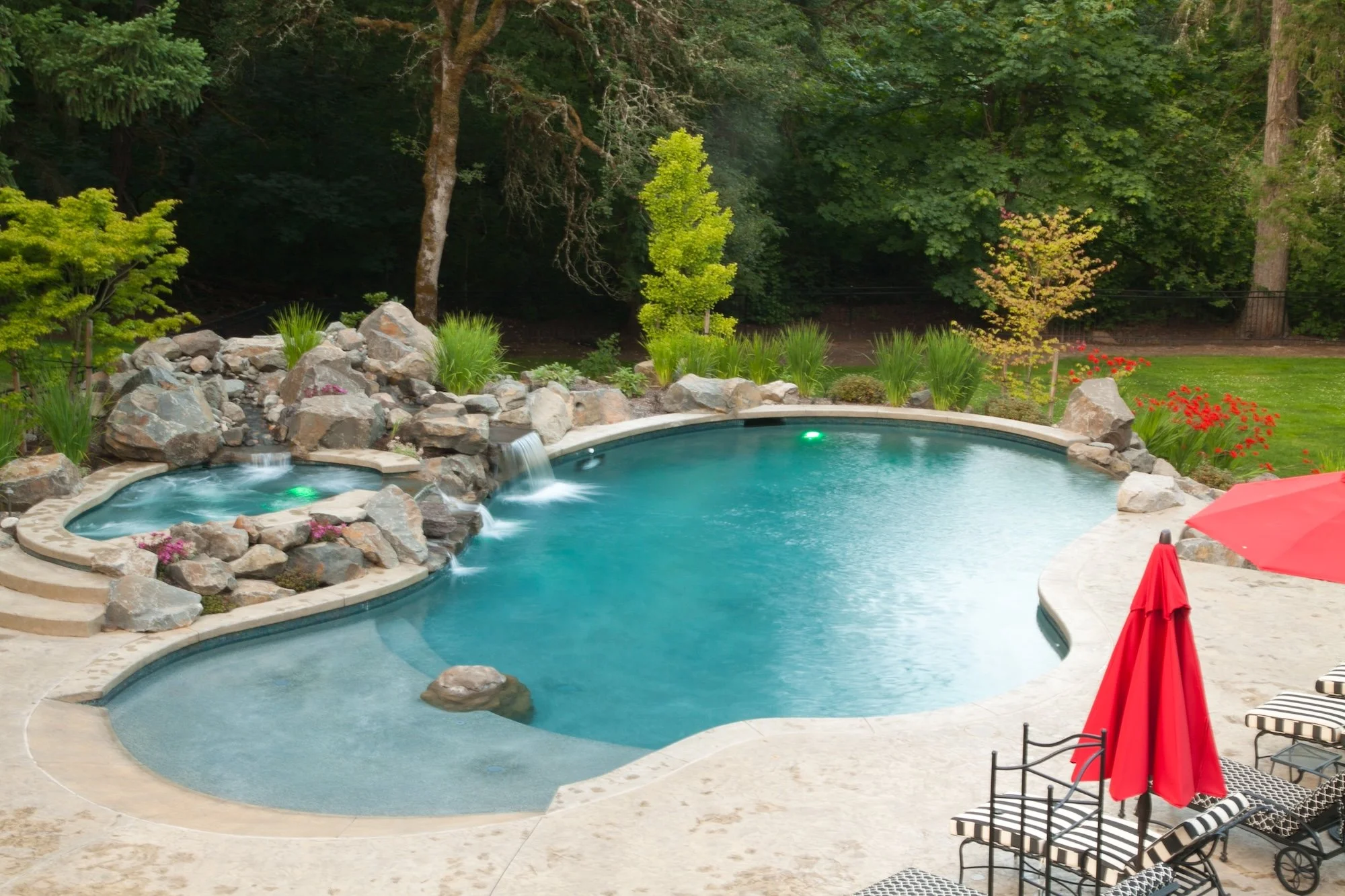 A backyard with a swimming pool, hot tub, and waterfall feature surrounded by rocks, plants, and trees. There are red umbrellas and striped chairs nearby.