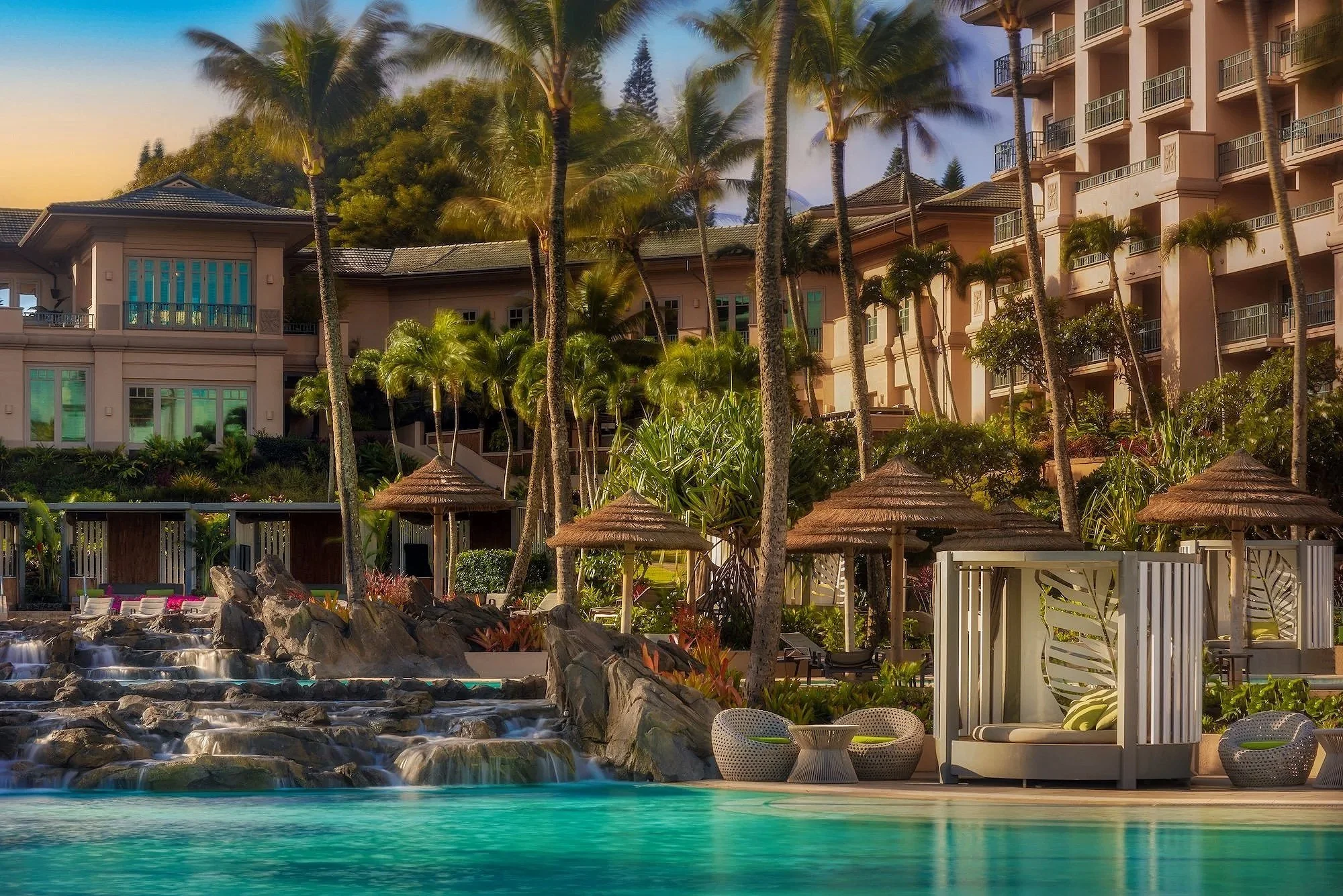Luxury resort with a large outdoor swimming pool, waterfalls, tropical landscaping, palm trees, umbrellas, sun loungers, and a multi-story building in the background during sunset.