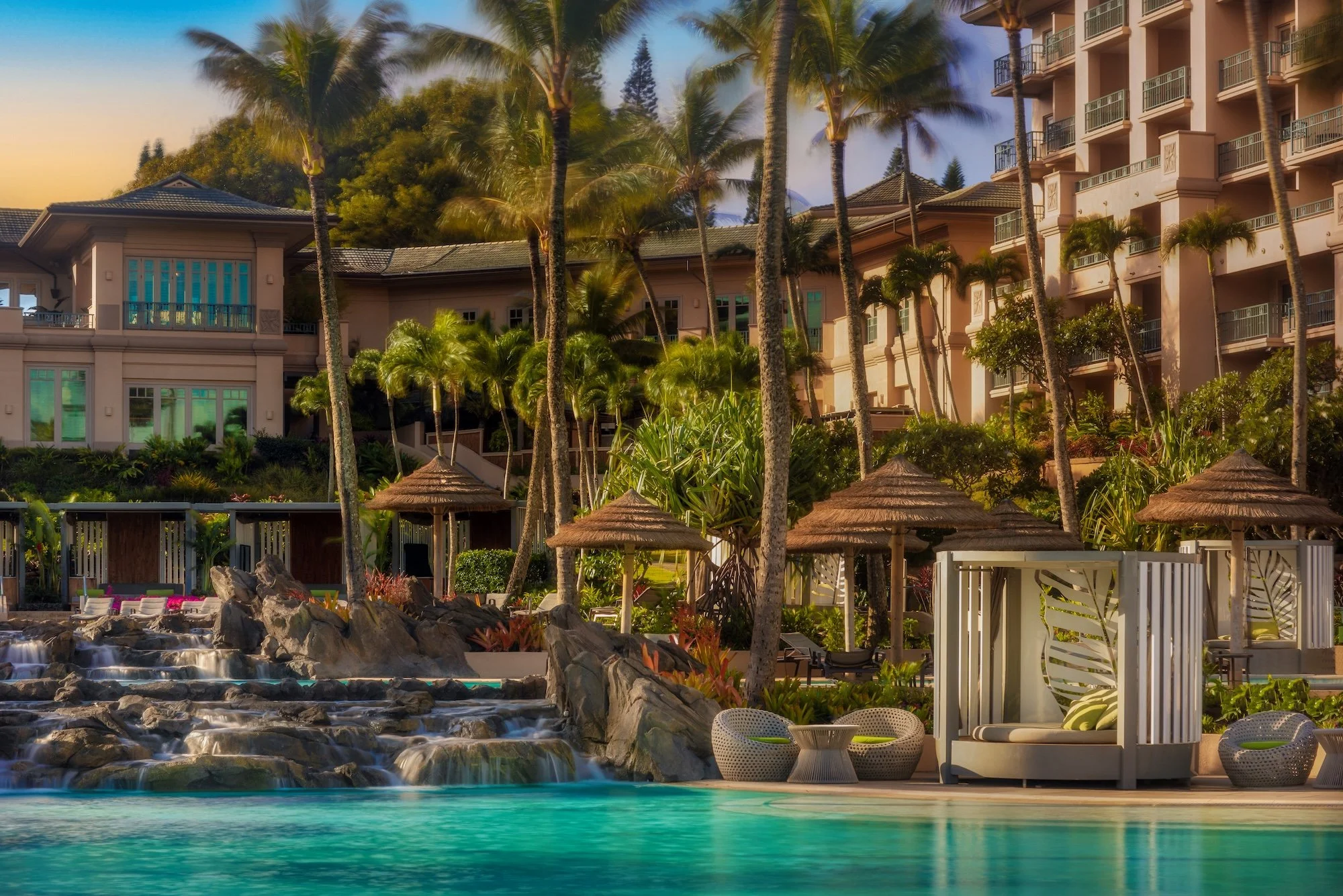View of a tropical resort with tall palm trees, a pink multi-story hotel building, waterfalls, and a swimming pool with poolside furniture and umbrellas.