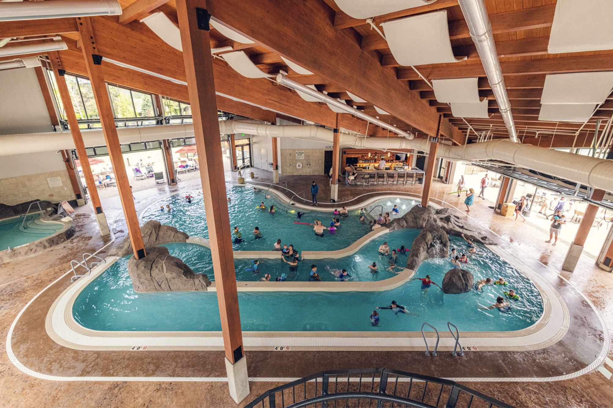 Indoor waterpark with a large swimming pool filled with children and adults, surrounded by rock formations, with a bar or seating area in the background. The area has large windows, wooden beams, and a high ceiling with exposed ducts and ventilation.