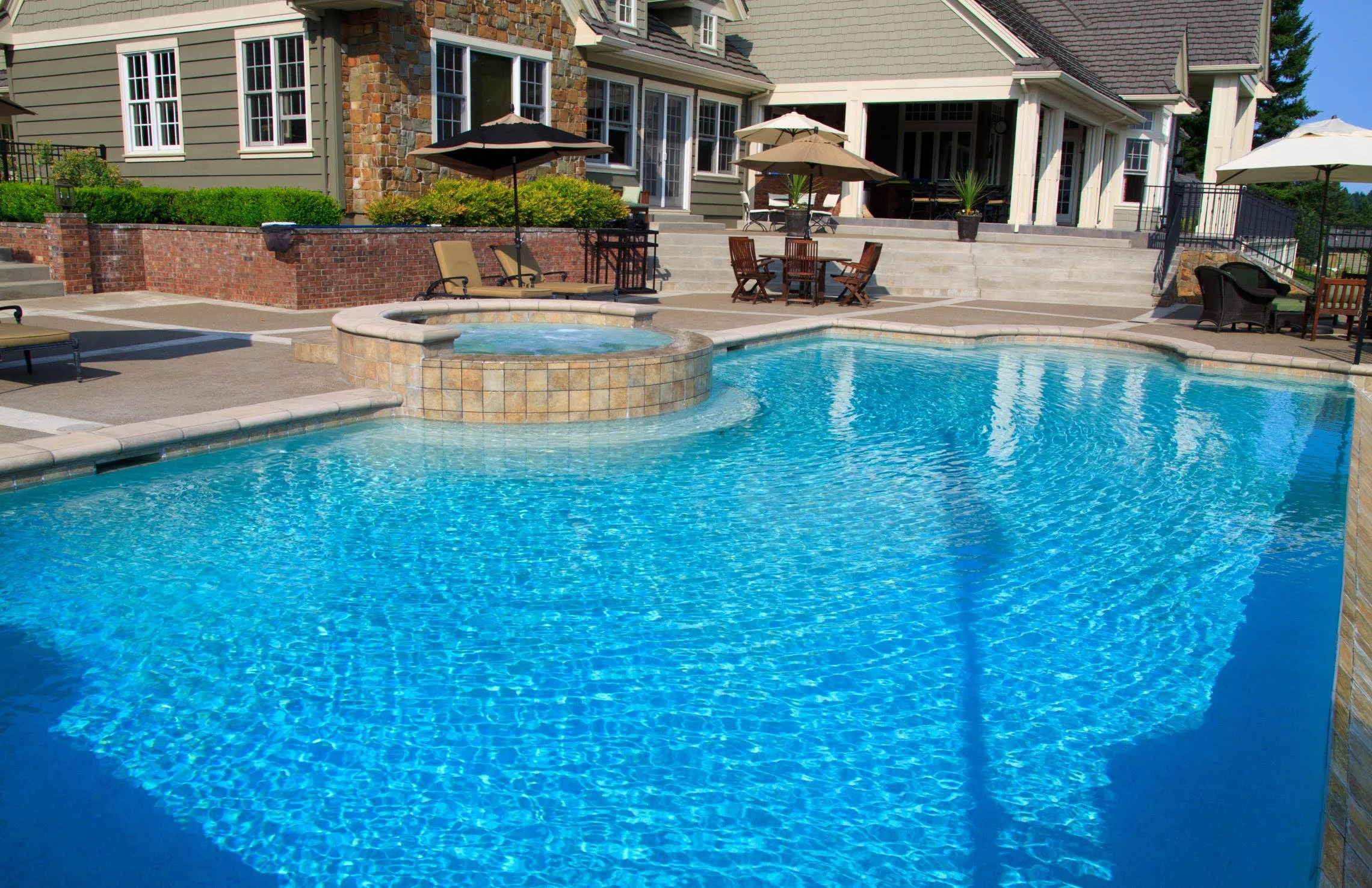 A backyard with a large swimming pool and hot tub, surrounded by outdoor furniture, umbrellas, and a house with multiple windows and a brick staircase.
