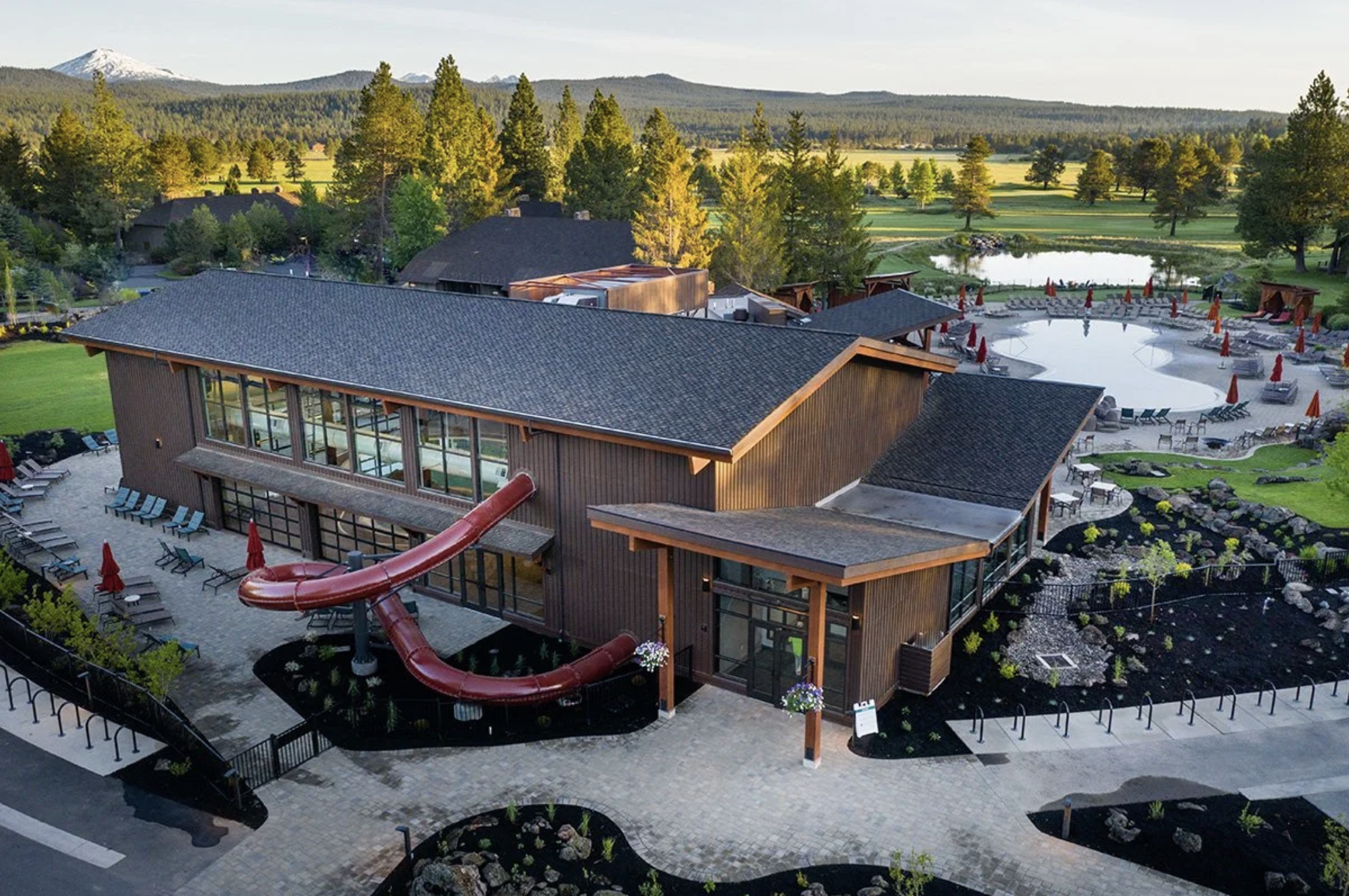A modern building with a dark gray roof and large windows is surrounded by a paved courtyard, lounge chairs, and a red water slide