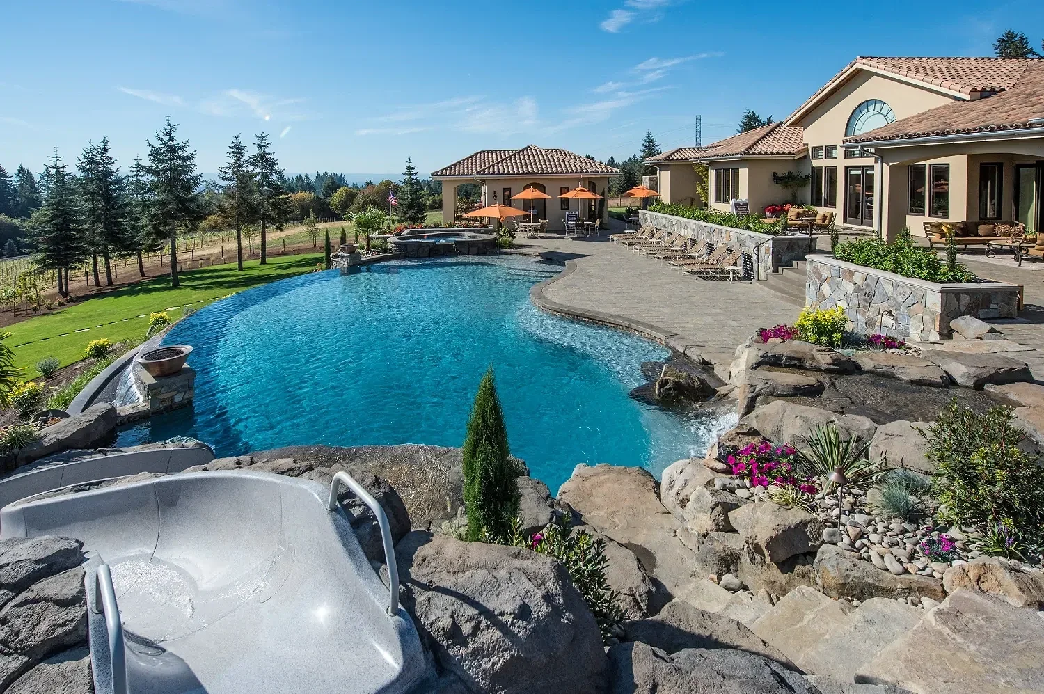 Residential backyard with an infinity pool, hot tub, landscaped garden, and a large house with a patio and lounge area under umbrellas, set against a scenic backdrop of trees and blue sky.