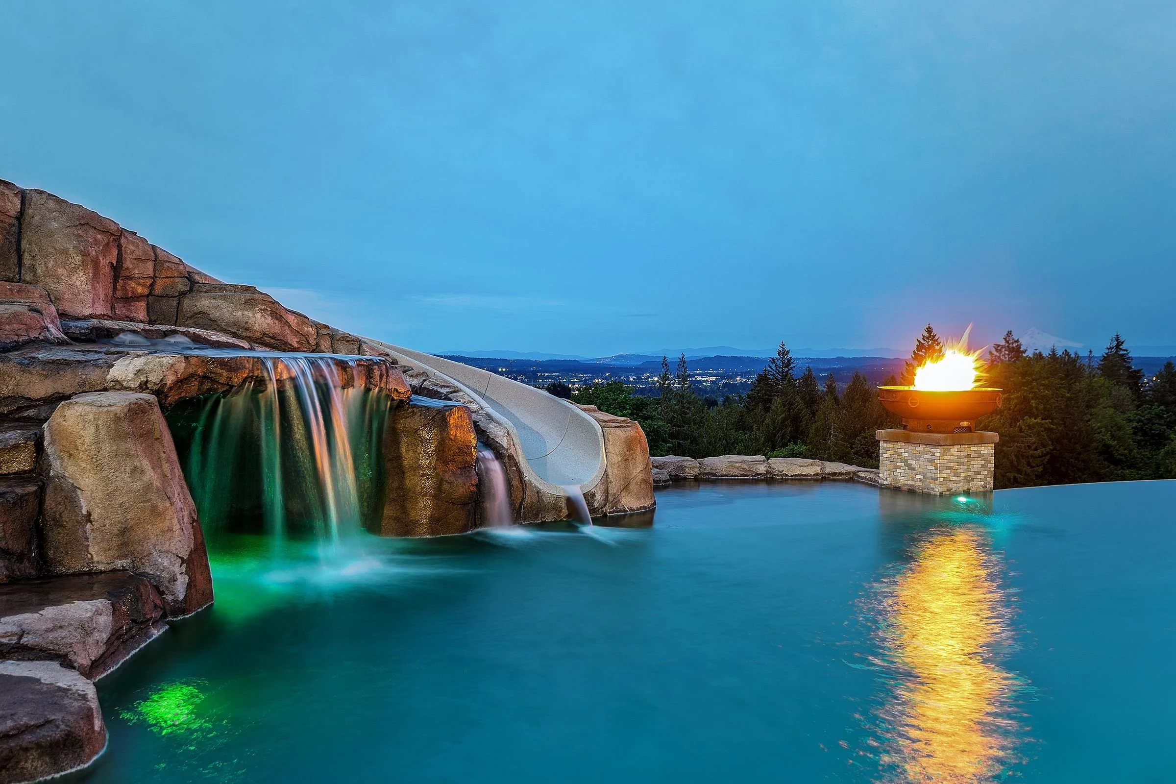 A scenic view of a waterfall cascading into a pool of water at sunset, with a fire pit on a stone base, trees, and hills in the background.