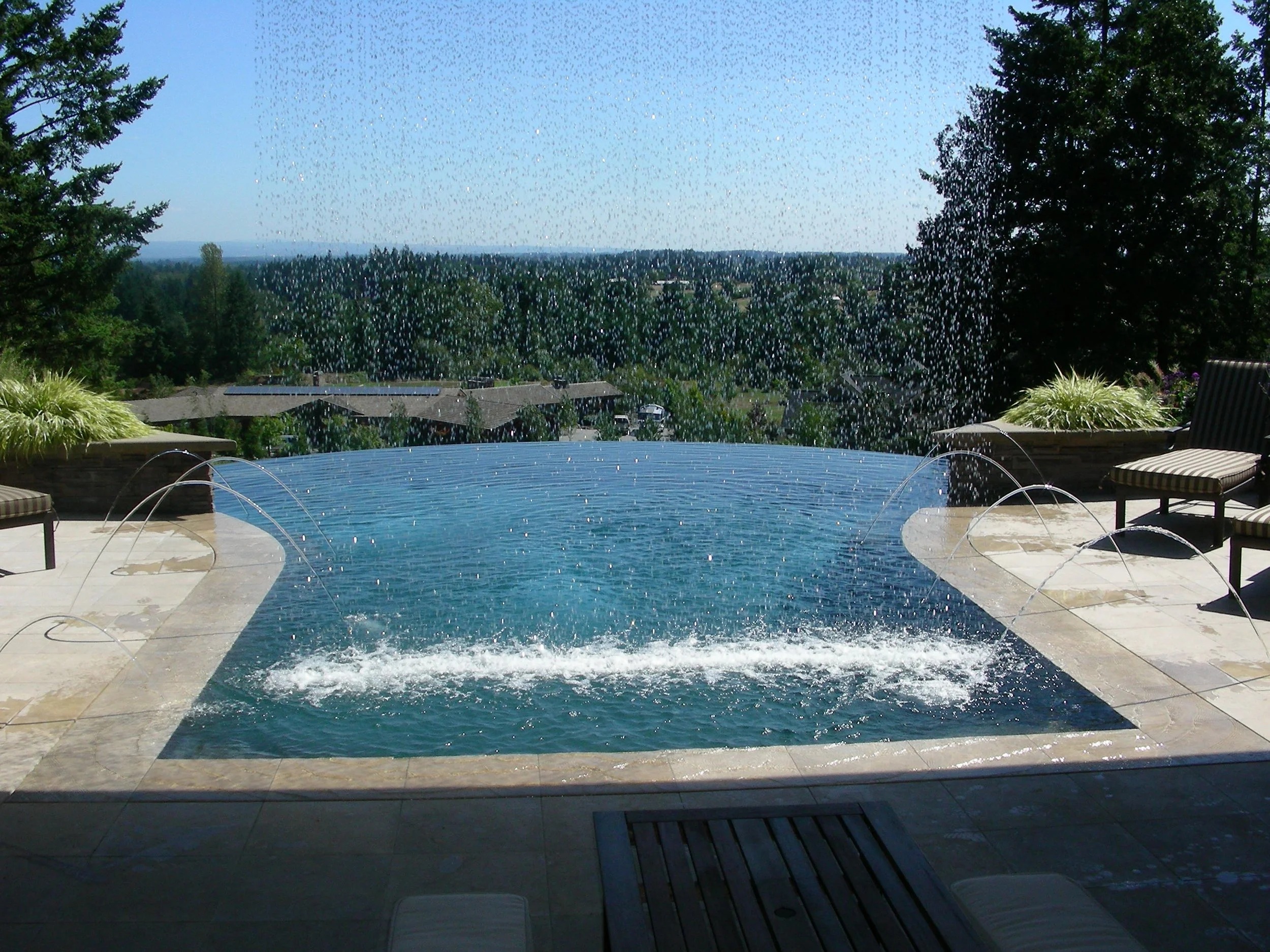 A backyard swimming pool with water fountains, surrounded by a deck and chairs, overlooking a forested landscape with houses and trees under a blue sky.