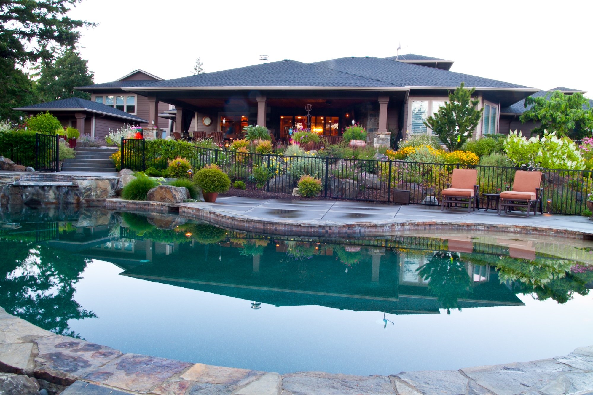 Large house with a spacious backyard featuring a swimming pool, surrounded by a variety of colorful flowers and greenery. There are patio chairs near the pool and the house has a deck area with outdoor furniture. The scene is during sunset.