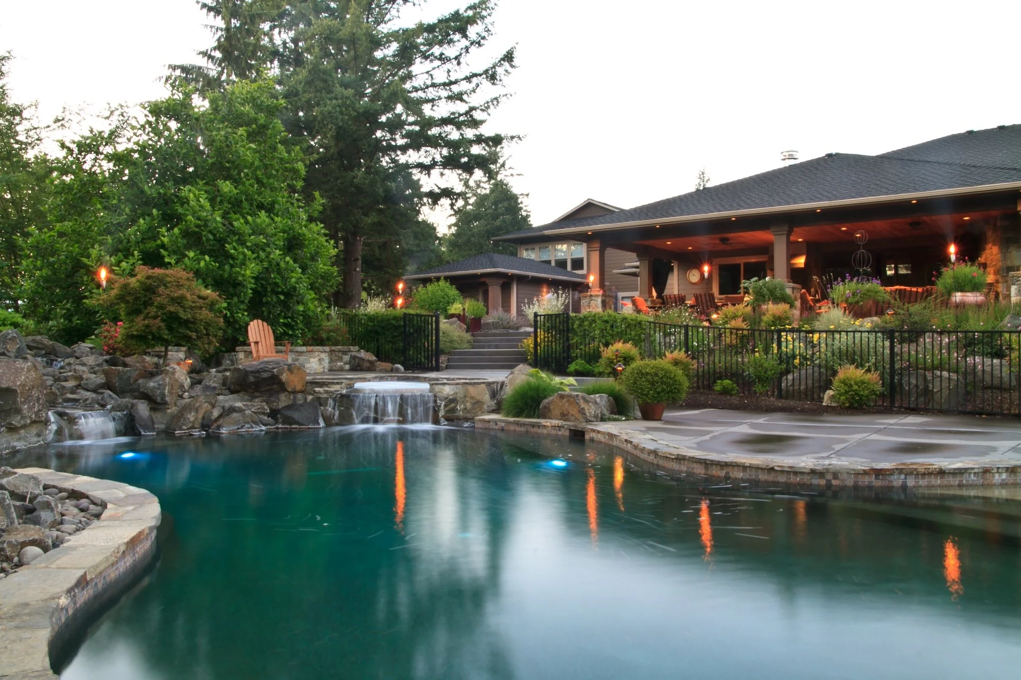 A backyard scene at dusk with a swimming pool, hot tub, lush greenery, and a house with a covered patio illuminated by outdoor lights.