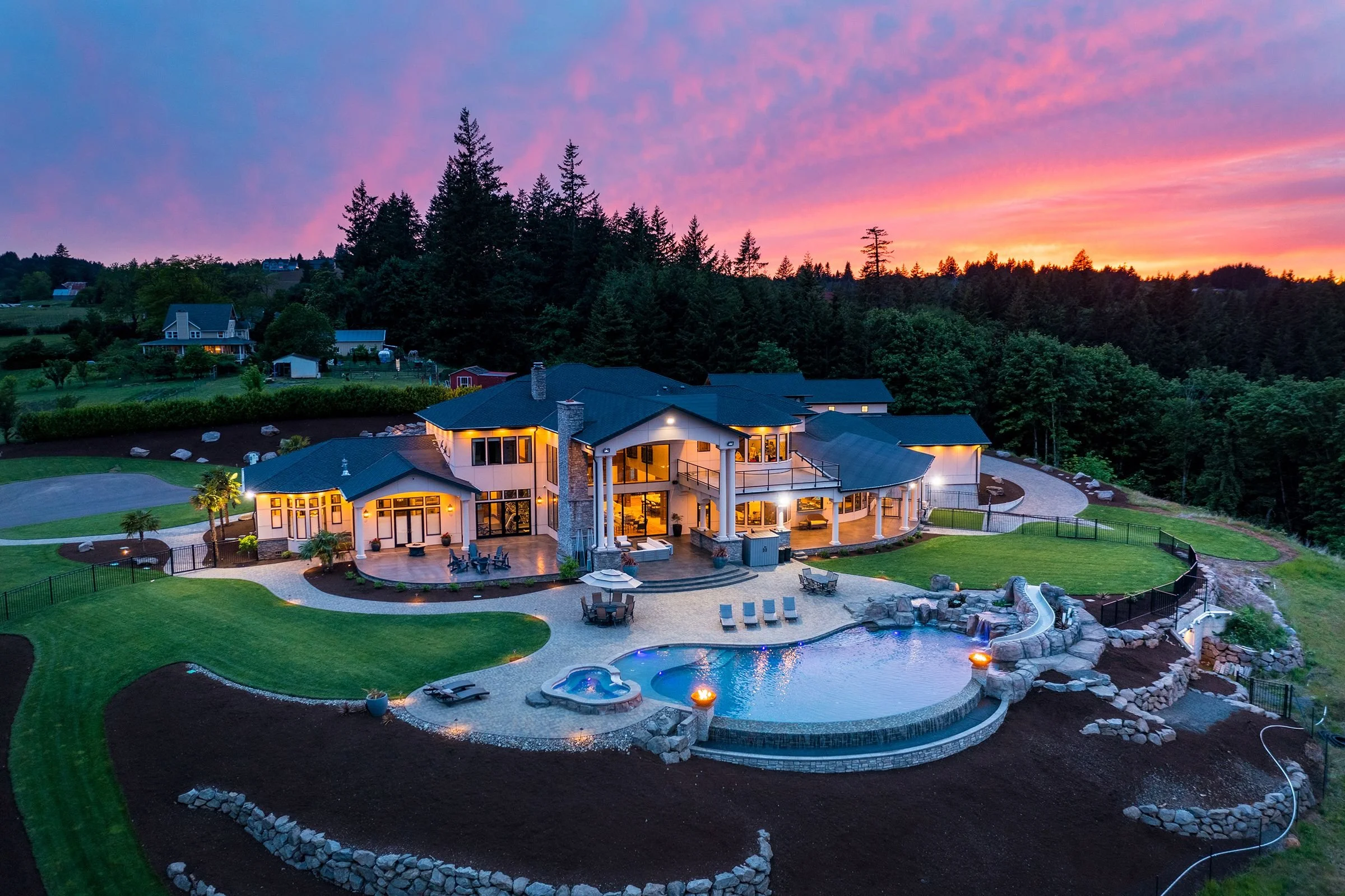 A large modern house with lit windows, a swimming pool, and a patio area at dusk, surrounded by lawns, trees, and neighboring houses on a hill, with a colorful sunset sky.