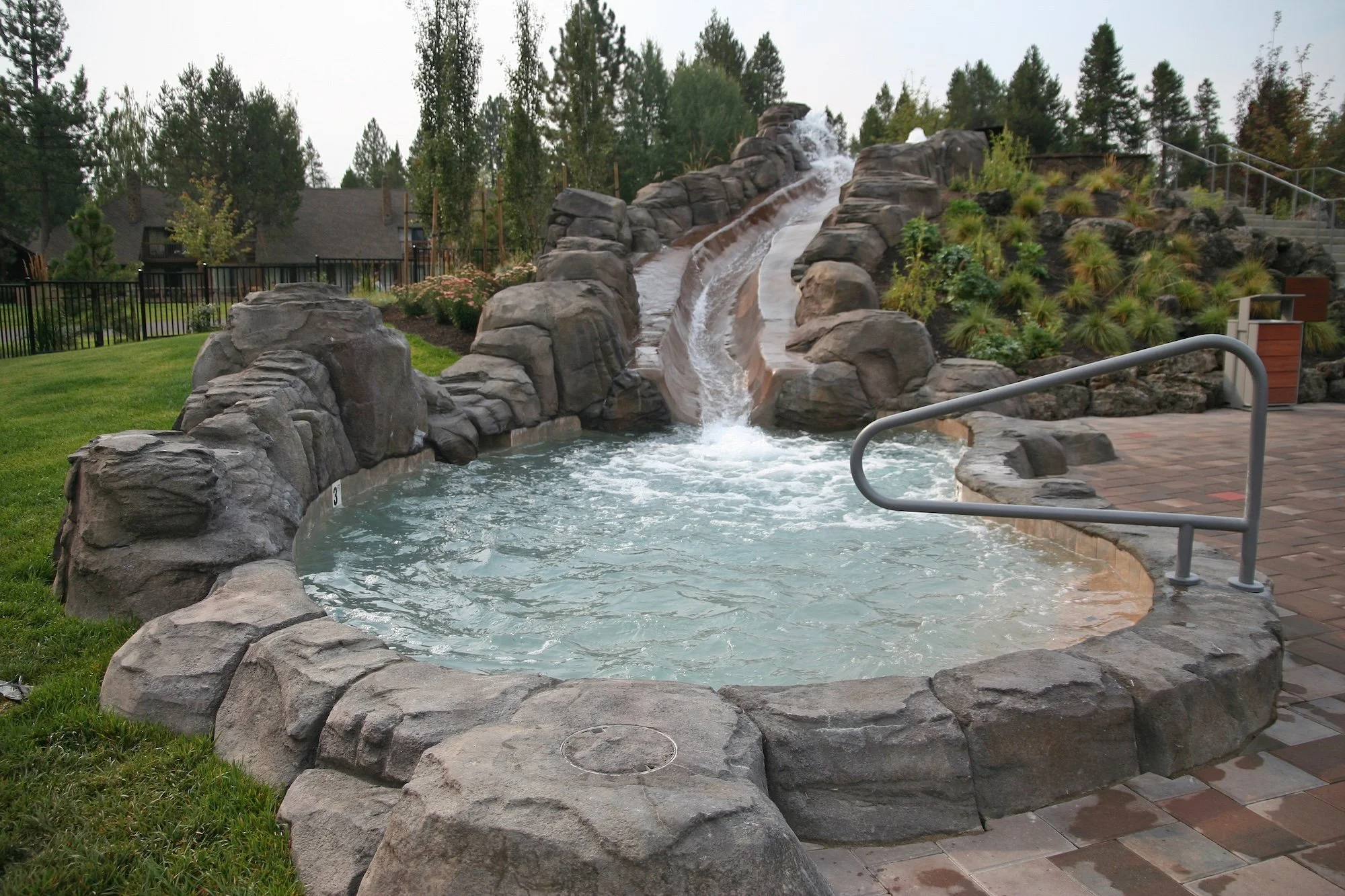 Outdoor hot tub with rock border and waterfall feature, surrounded by greenery and trees, with a residential building and fenced yard in the background.