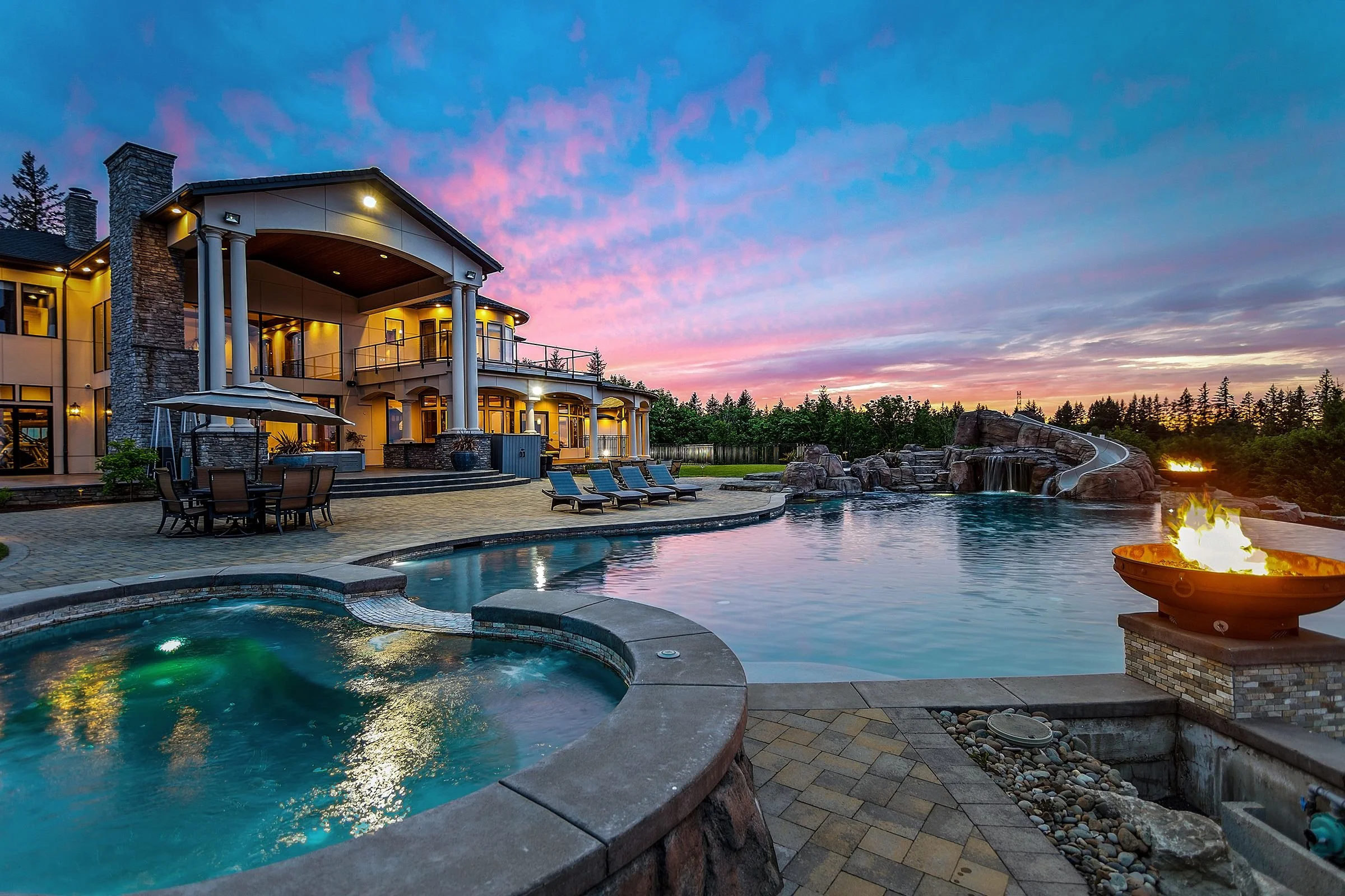 A luxurious house with a swimming pool and hot tub in the backyard during sunset, with lounge chairs and an umbrella around the pool.