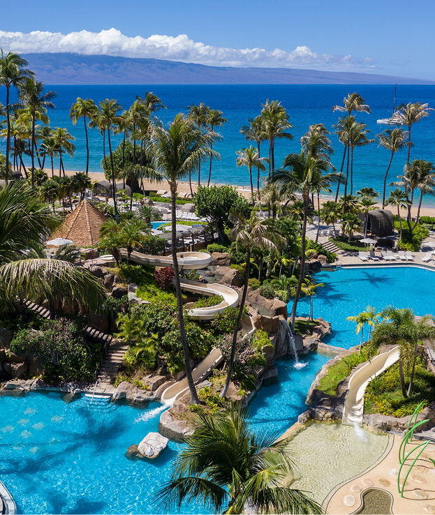 A view of a tropical resort with swimming pools, palm trees, and a beach with ocean in the background.