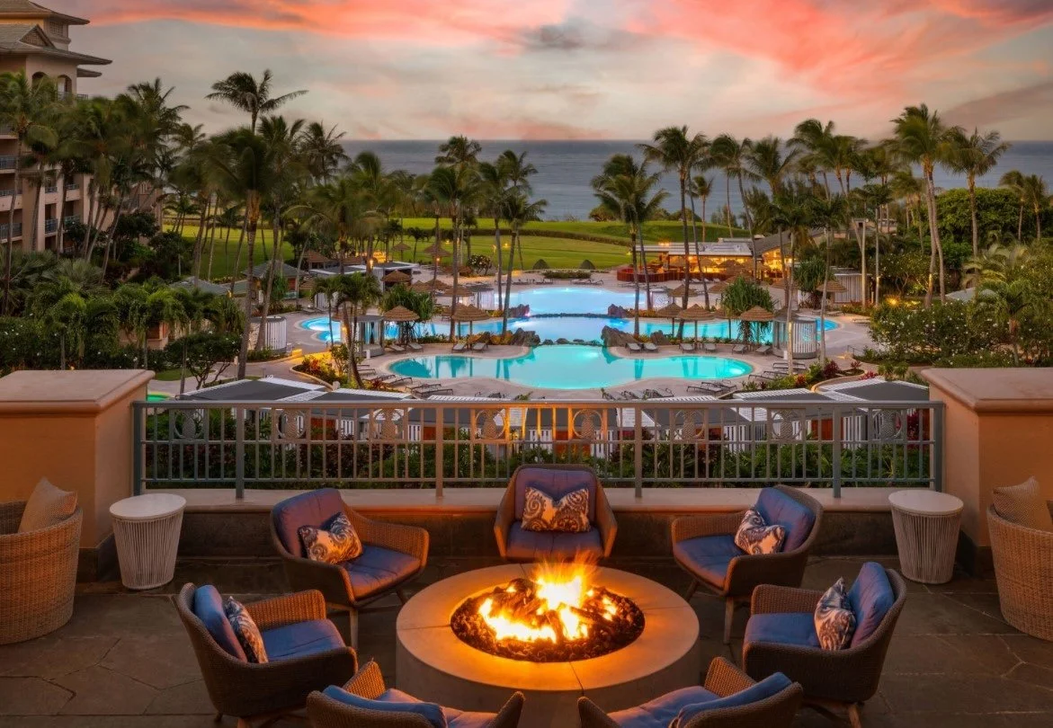 A luxurious outdoor pool area at sunset, overlooking the ocean with palm trees, lounge chairs, and a fire pit surrounded by chairs.