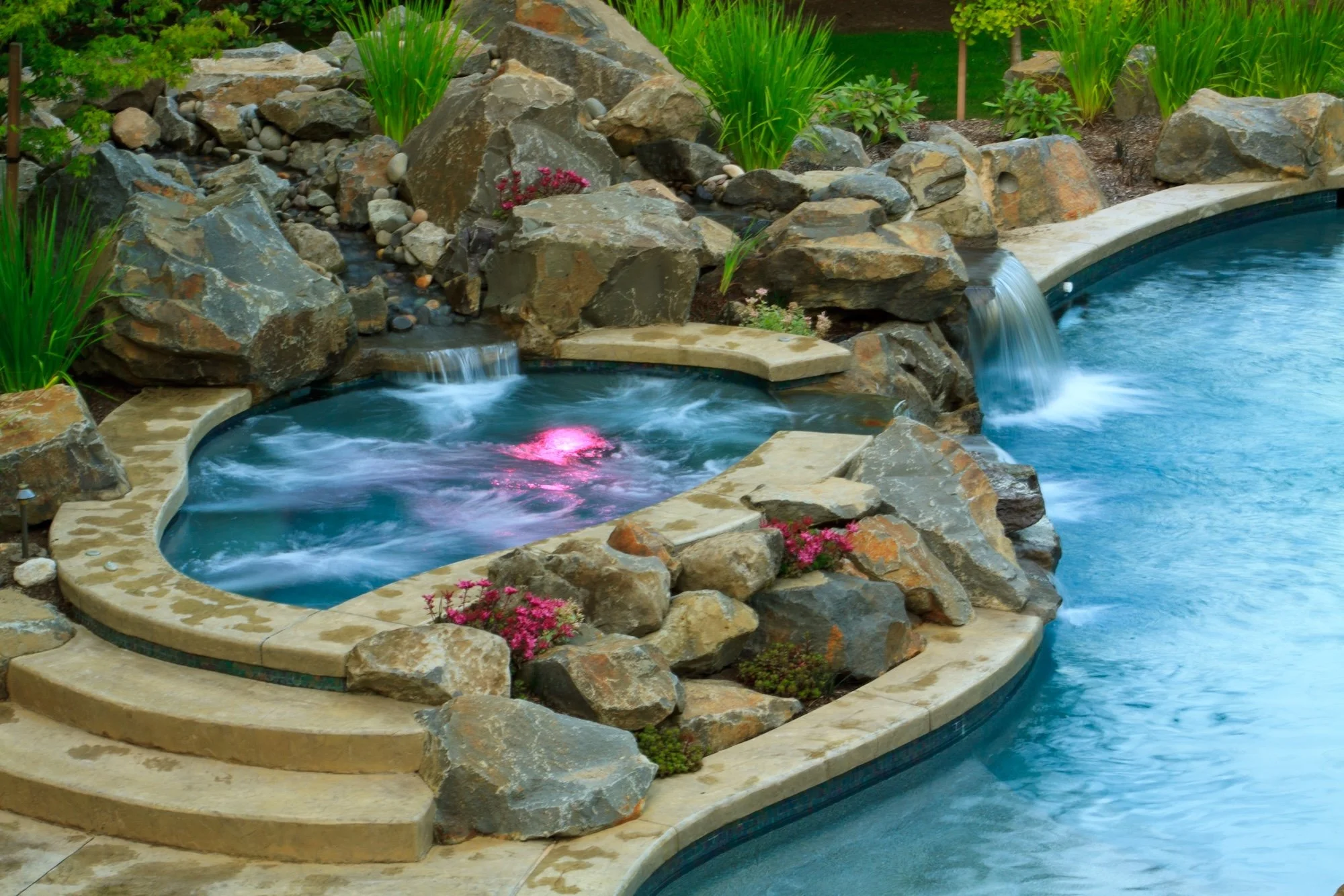 Resort swimming pool with a hot tub, rocks, and a water feature in a landscaped yard.