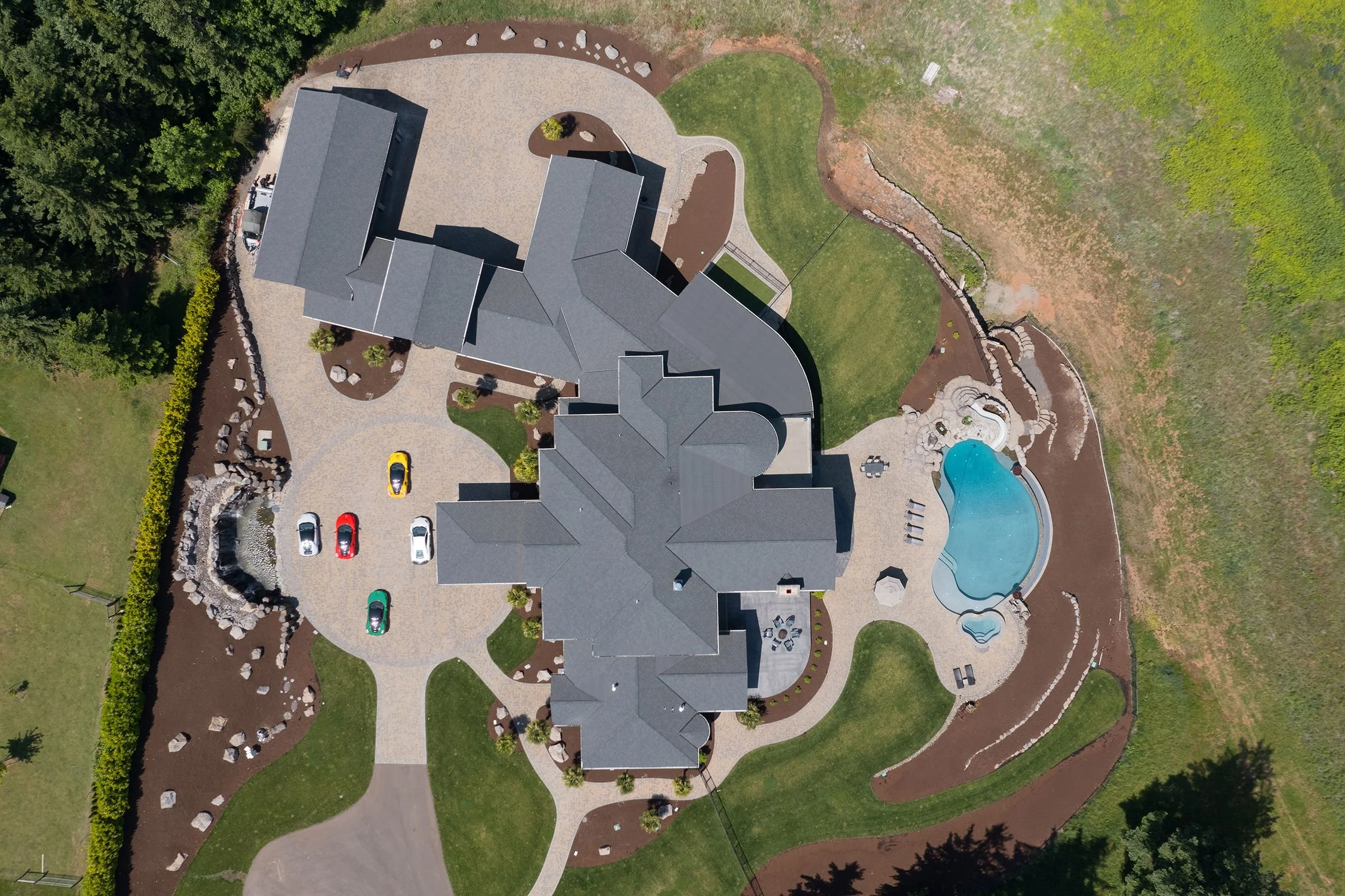 Aerial view of a large house with a swimming pool, surrounding driveway with parked cars, landscaped yard, and trees.