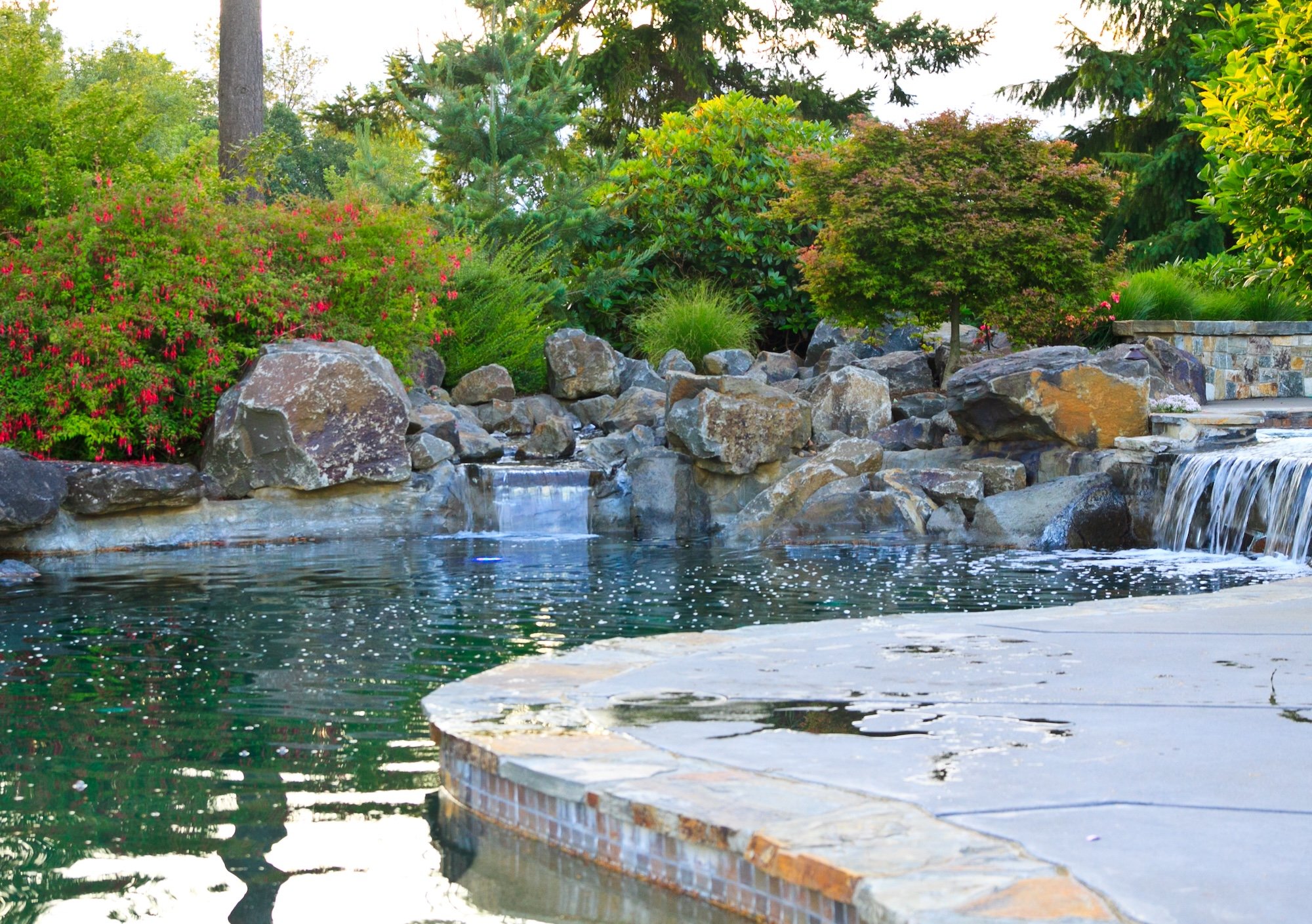 A small waterfall flowing over rocks into a pond, surrounded by lush green trees and vibrant shrubs in a landscaped garden setting.