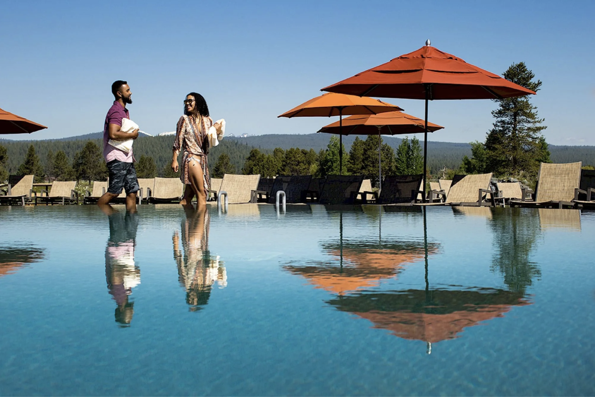 A man and woman are walking in a swimming pool, holding towels, with a scenic outdoor setting in the background. There are several orange and brown umbrellas along the poolside, and lush green trees with mountains in the distance under a clear blue sky.