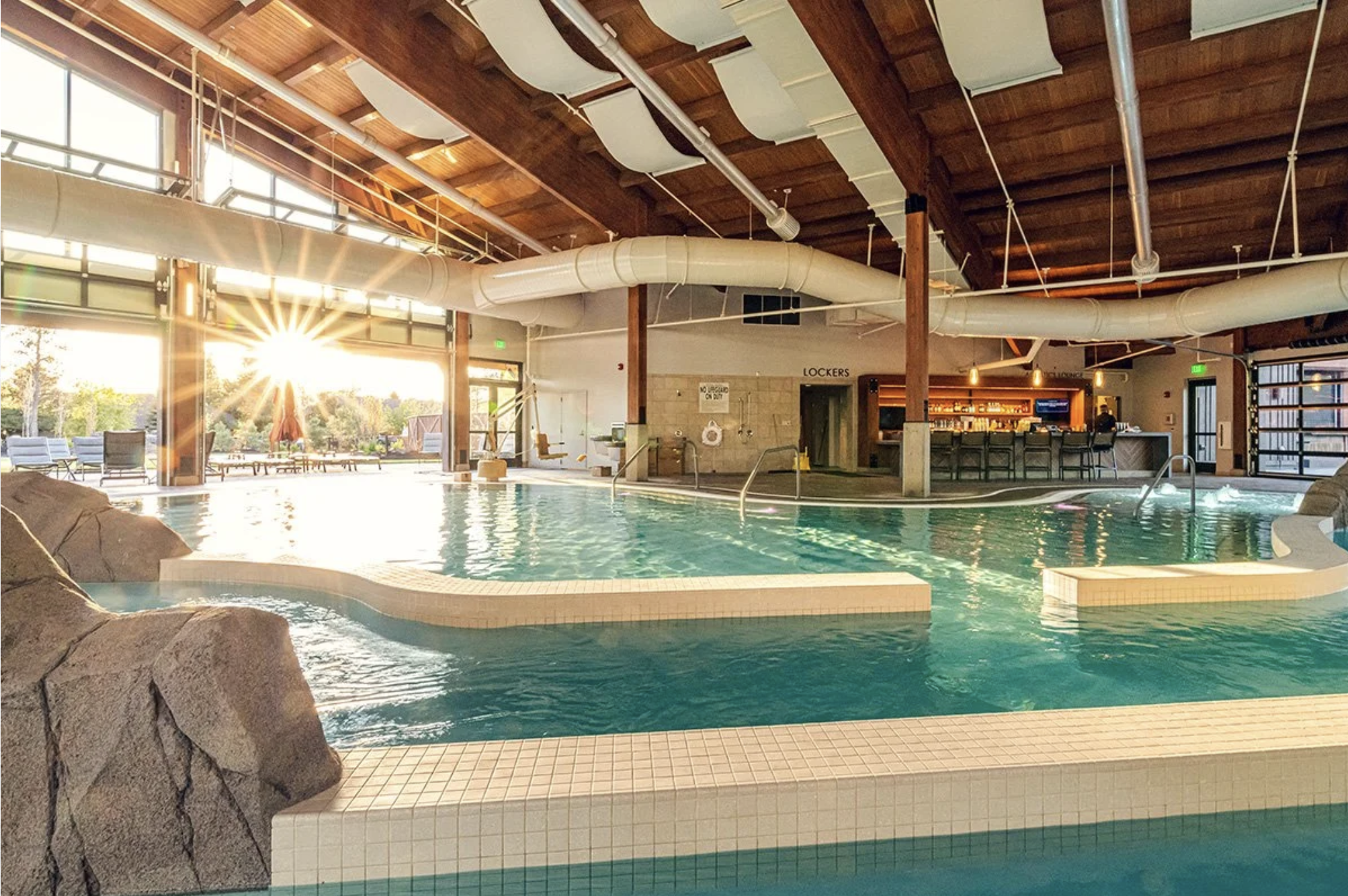 Indoor swimming pool area with large windows, water slide, lounge chairs, and a bar area in the background