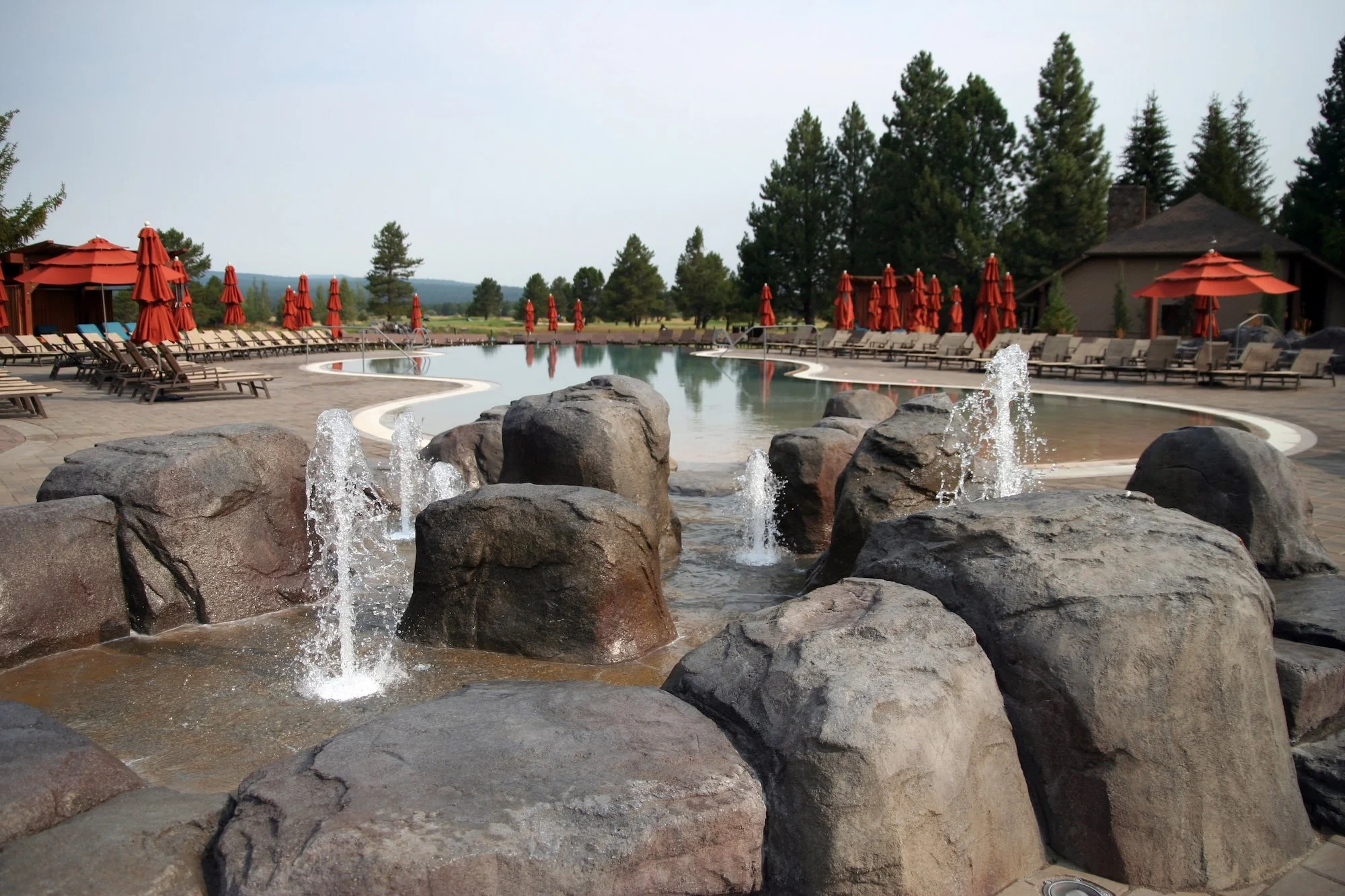 A large outdoor swimming pool surrounded by lounge chairs and red umbrellas, with a stone fountain in the foreground and trees in the background.