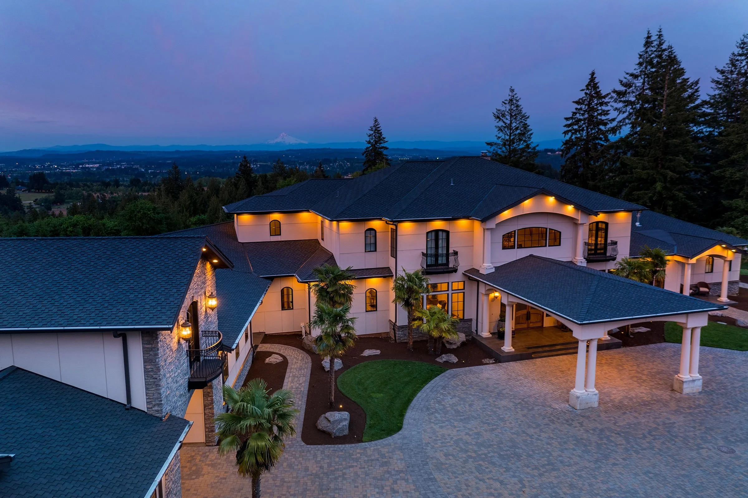 Aerial view of a luxurious house with a courtyard and trees, overlooking a cityscape with a mountain in the distance during dusk.