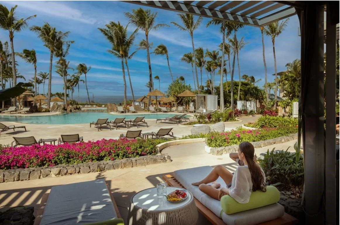 A woman relaxing on a lounge chair by a resort pool with palm trees and blue sky in the background.