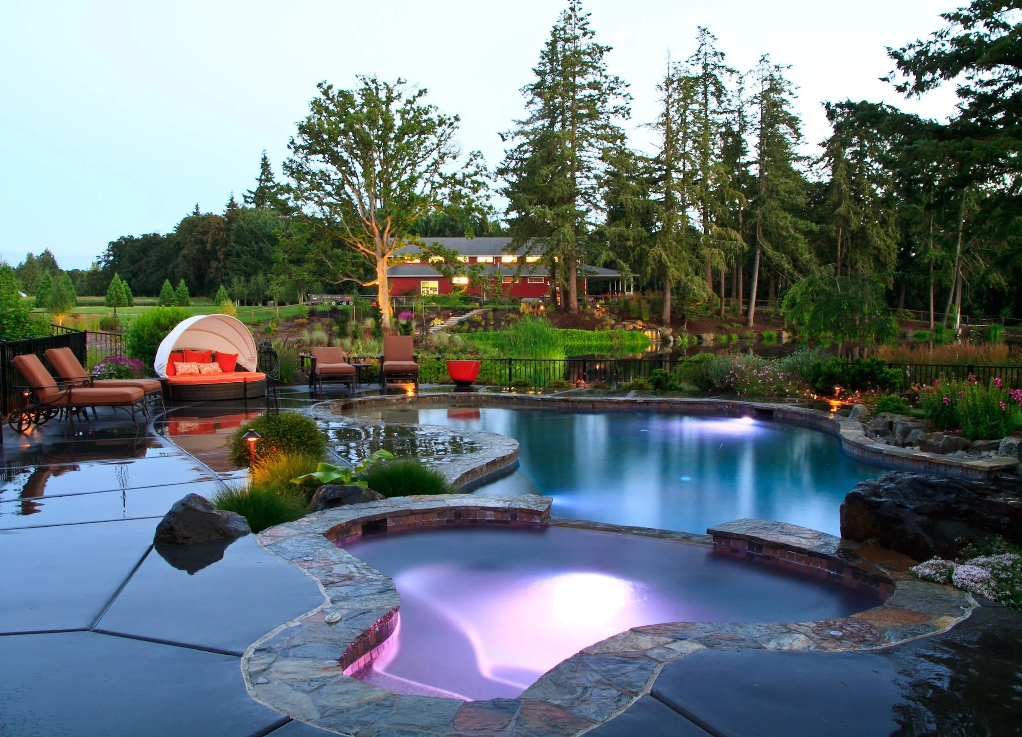 A backyard with a swimming pool surrounded by rocks and furniture, lush greenery, tall trees, and a red house in the background during evening with lighting.
