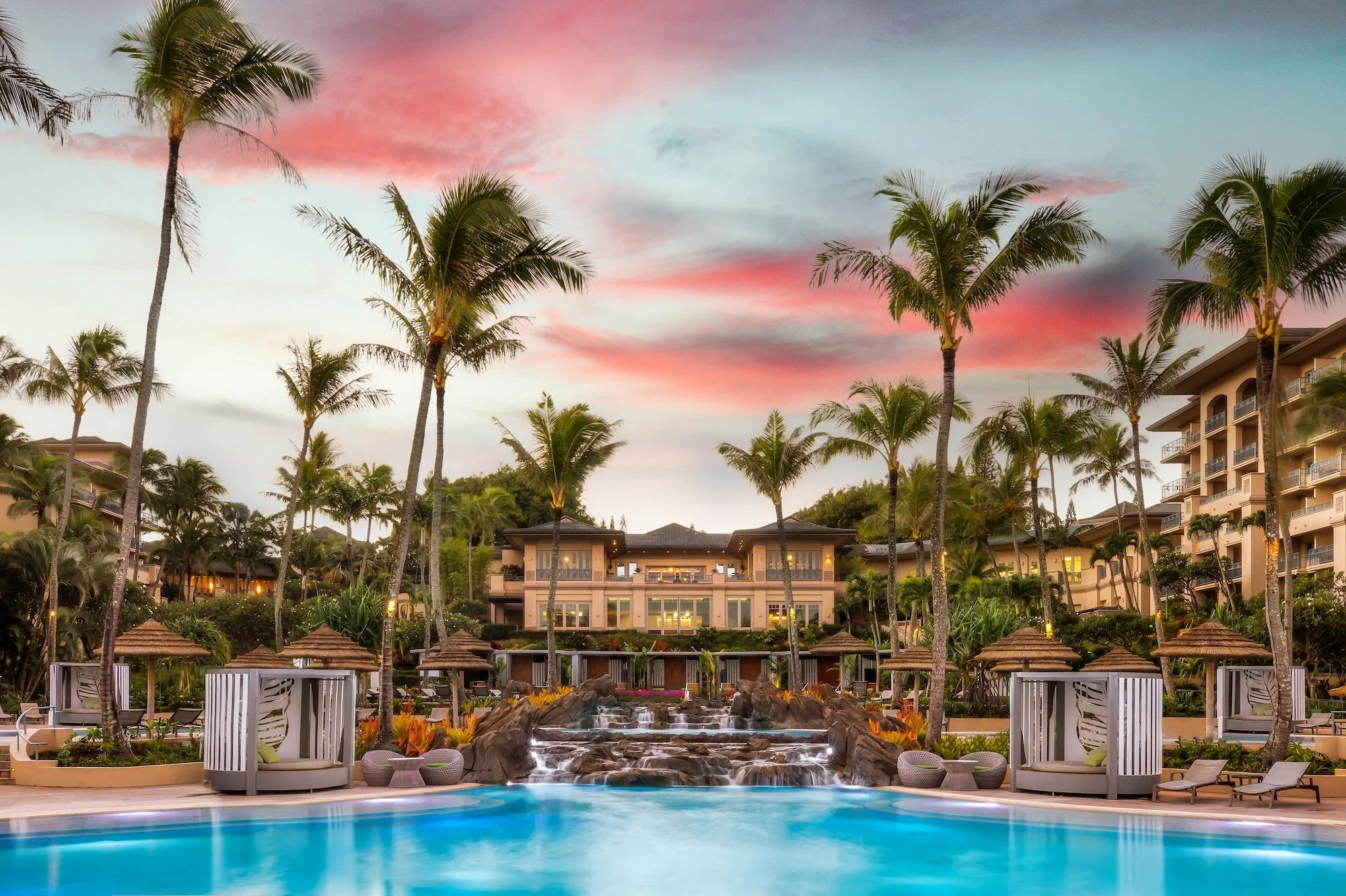 Resort with a large swimming pool, waterfall feature, lounge chairs, and cabanas, surrounded by palm trees under a colorful sunset sky with pink and blue hues, and a multi-story building with balconies in the background.