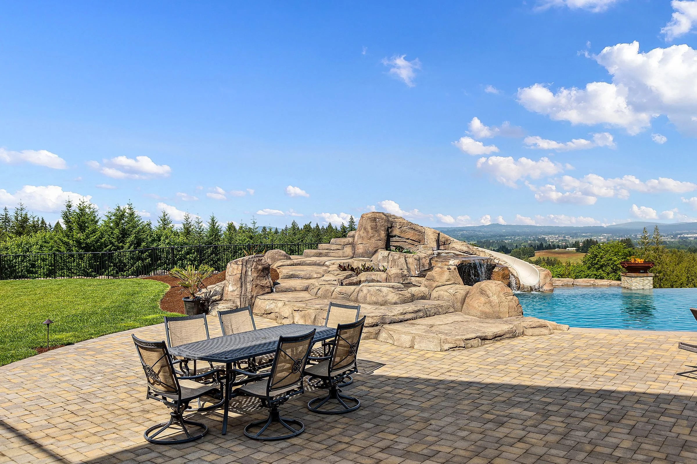 Outdoor patio with black metal table and chairs, stone waterfall feature, swimming pool, lush green lawn, and a scenic view of trees and distant mountains under a blue sky with scattered clouds.
