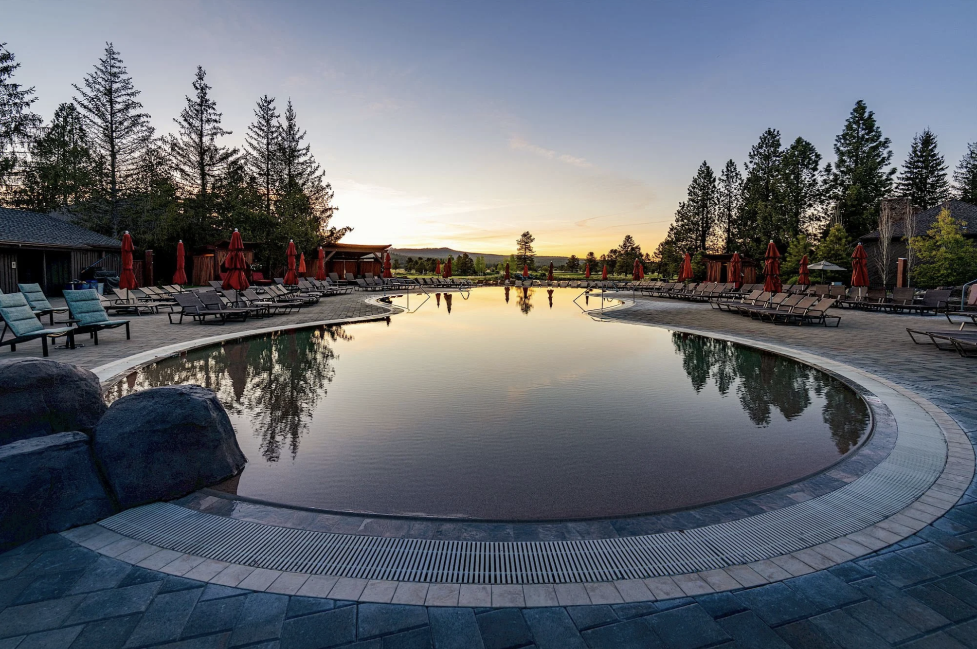 An outdoor swimming pool area with lounge chairs and red umbrellas, surrounded by trees, at sunset.