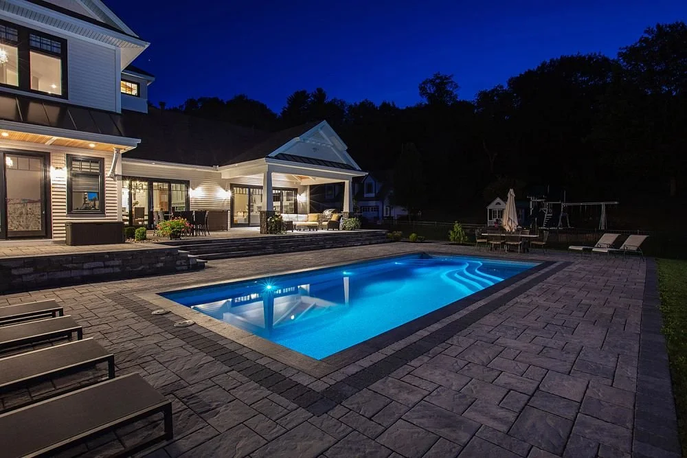 Nighttime view of a backyard with a lit rectangular swimming pool, patio furniture, and a modern house with large windows and exterior lighting.