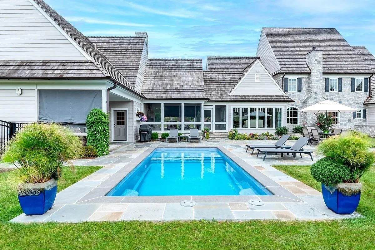 Backyard with a rectangular swimming pool, lounge chairs, patio furniture, potted plants, and a house with large windows and a stone chimney.
