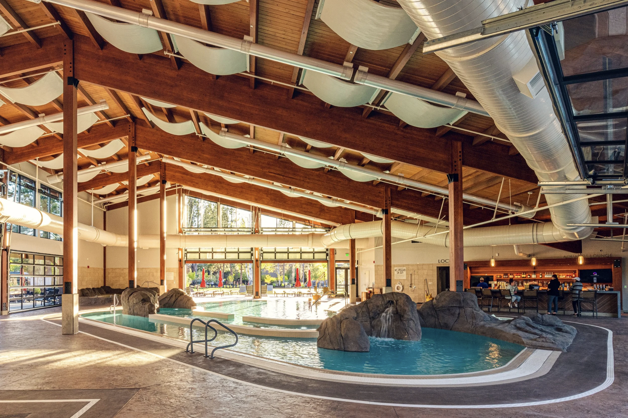 Indoor swimming pool area with a wooden ceiling, large windows, and outdoor seating visible through the windows.