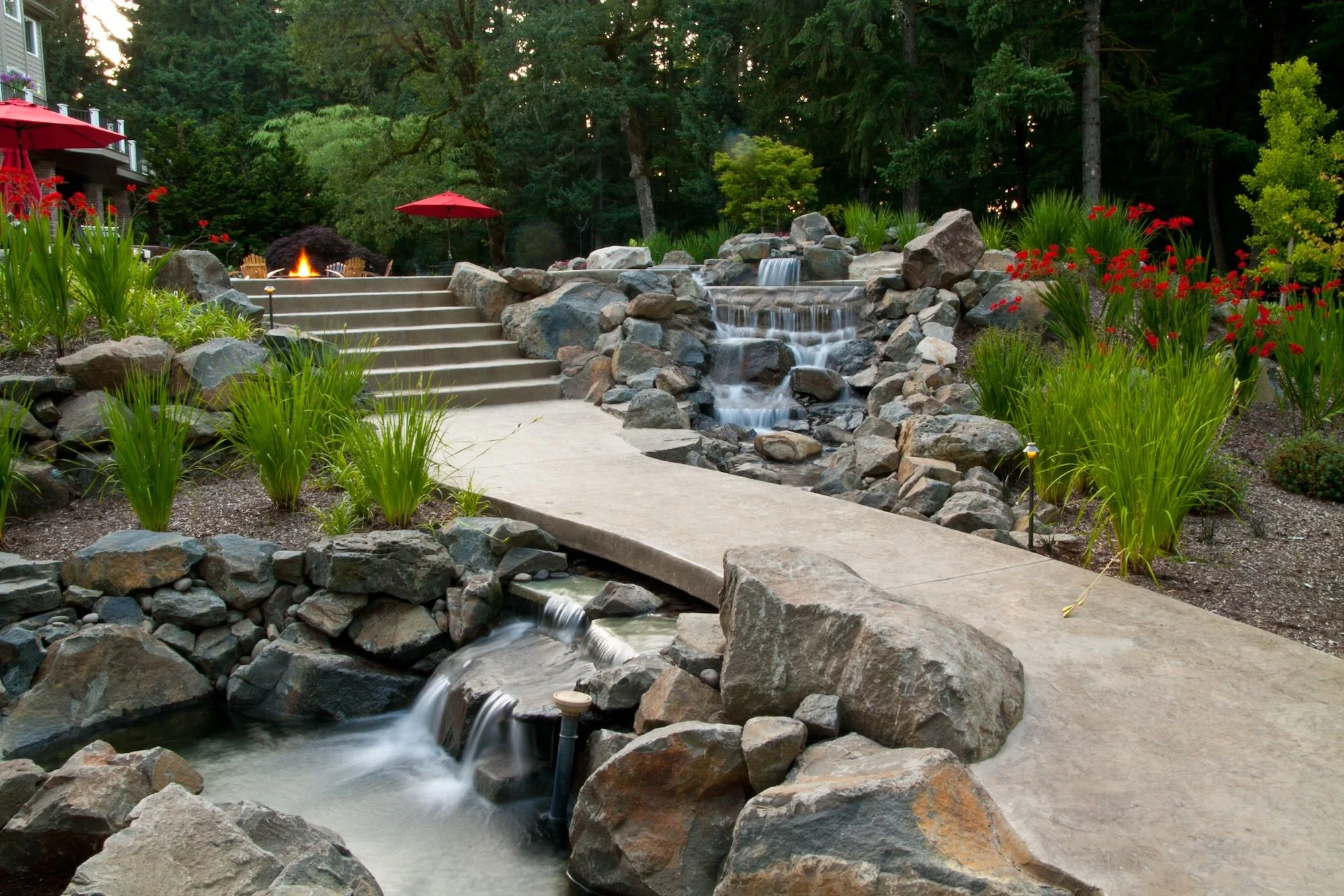A backyard landscape featuring a waterfall with multiple tiers flowing over rocks, a concrete pathway, and a fire pit area with red umbrellas. Surrounding the area are lush green plants and tall trees, creating a serene outdoor space.