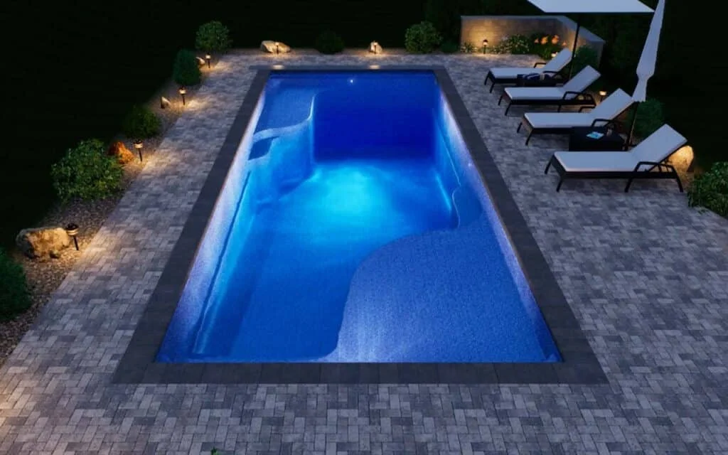A lit swimming pool at night surrounded by a paved patio area with lounge chairs and greenery.