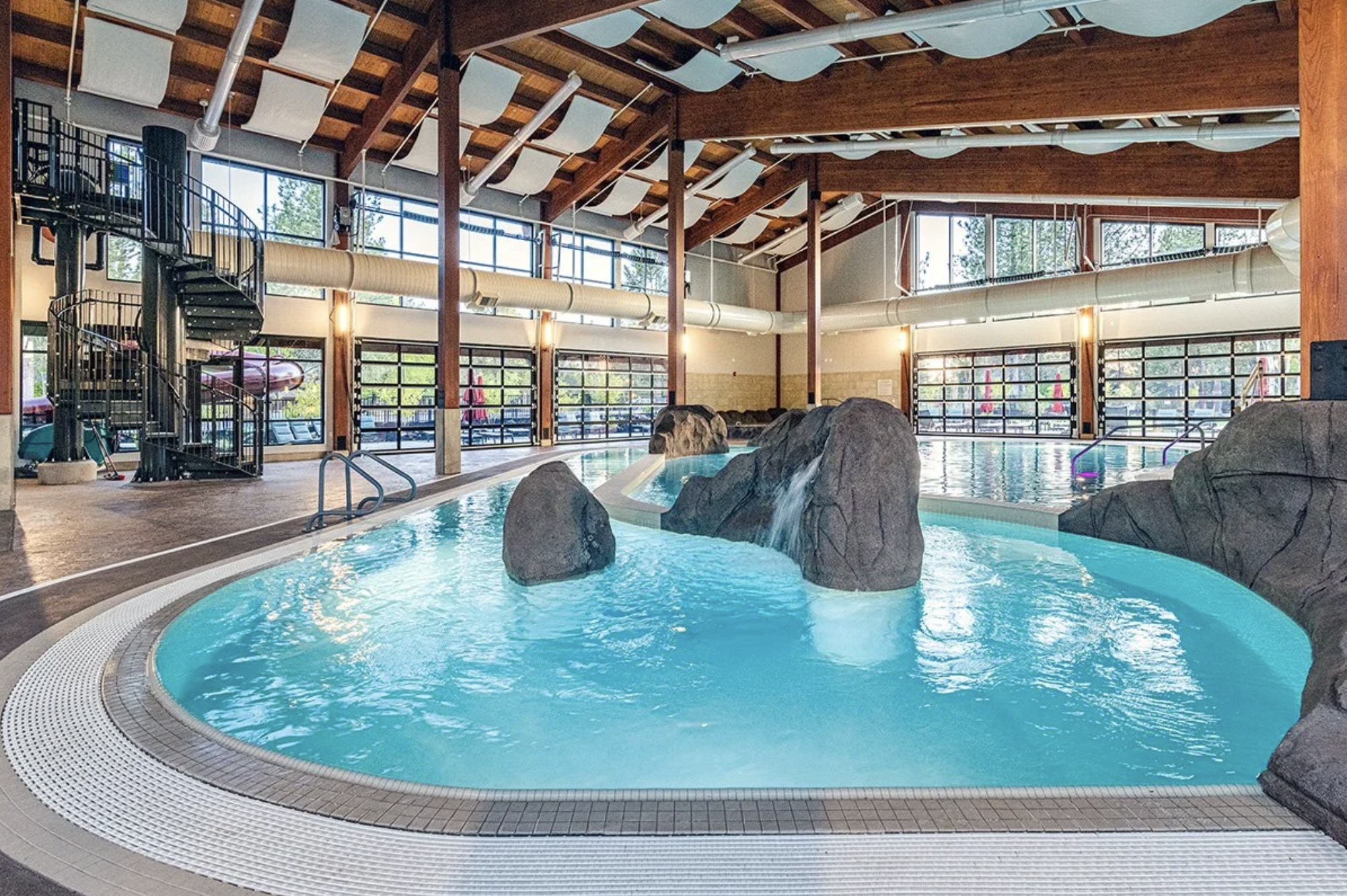 Indoor swimming pool with a rock waterfall feature and a large hot tub, surrounded by floor-to-ceiling windows and wooden beams ceiling.