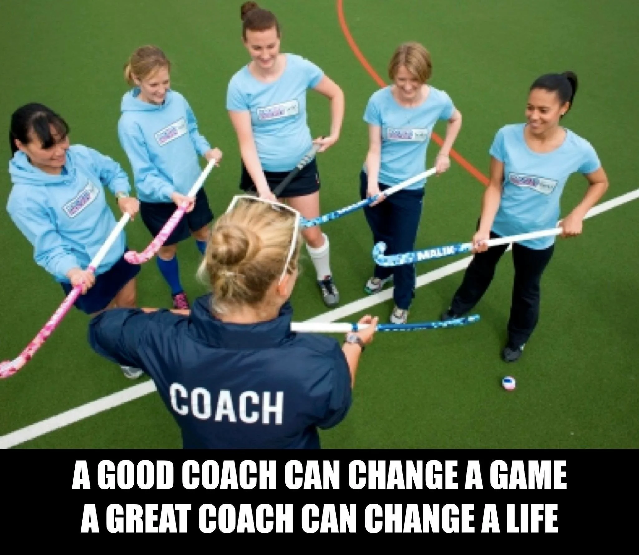 A coach with 'COACH' printed on the back, standing in front of a group of five female field hockey players holding sticks, on an indoor field. The players are listening to the coach during a training session.