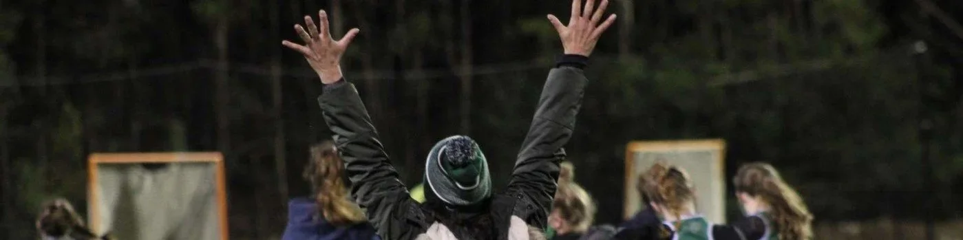 A person with a beanie raising their arms in celebration on a field with people in the background.