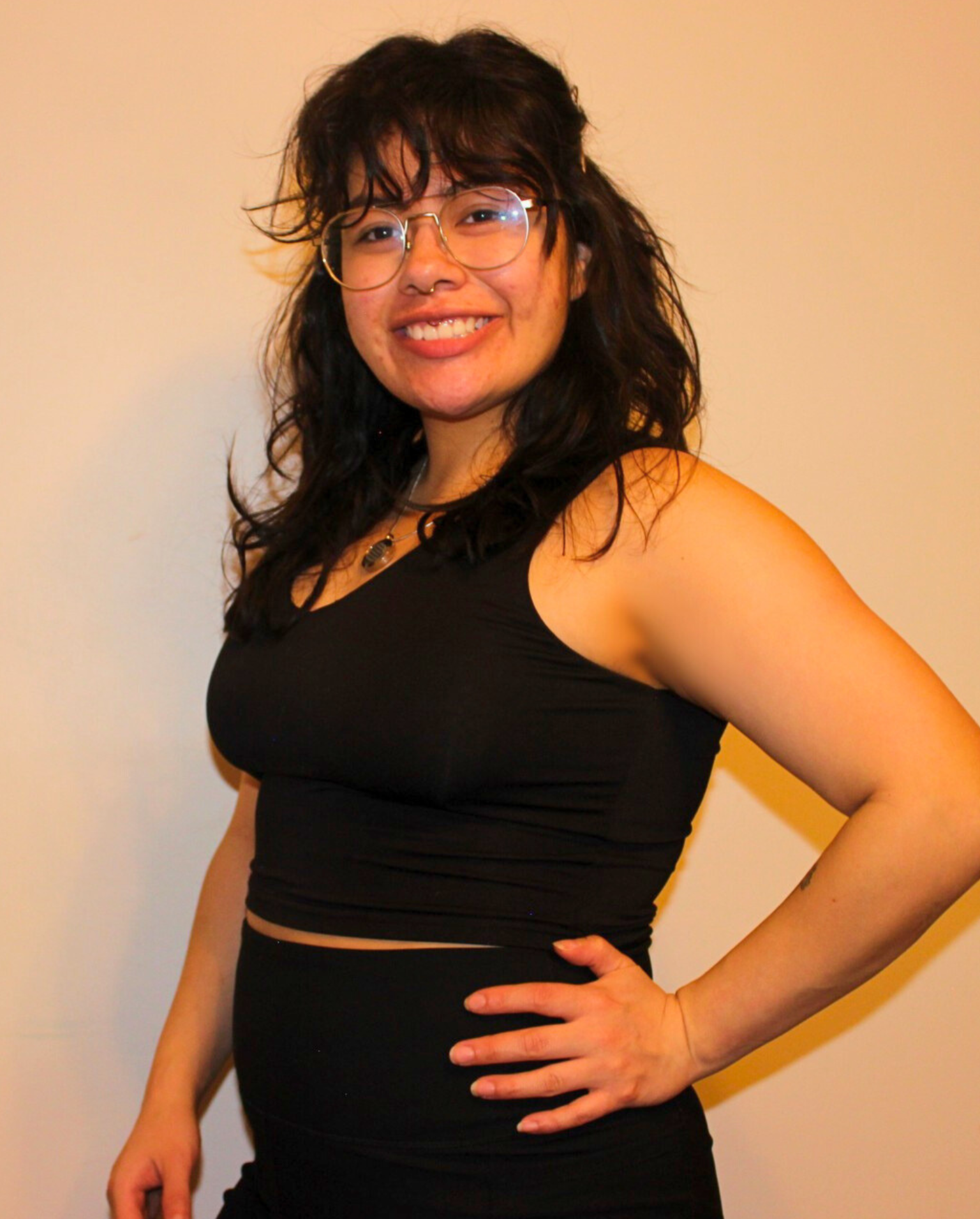 Young woman wearing glasses and a black tank top, smiling and posing with one hand on her hip against a plain wall.