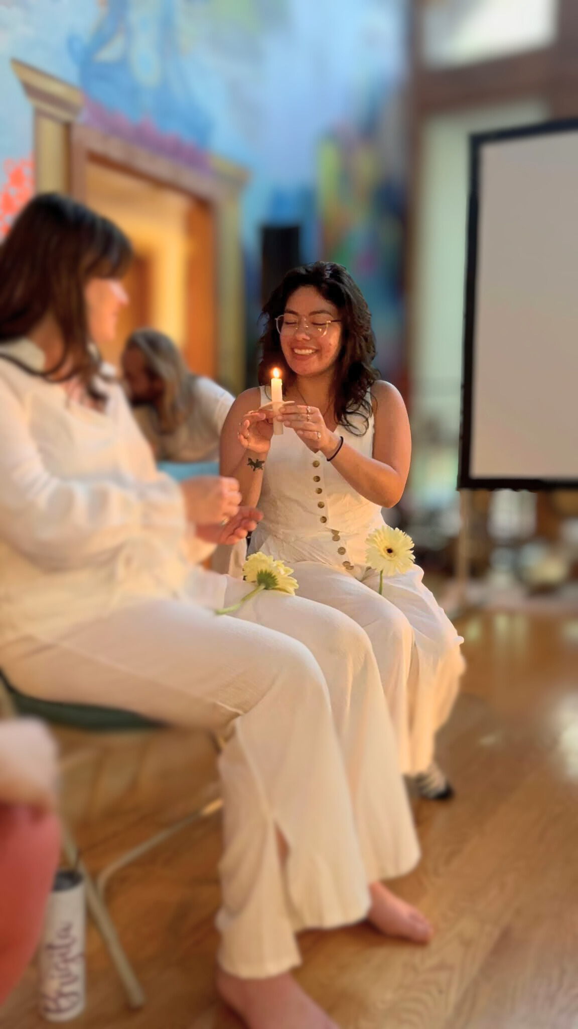 Two women in white dresses participating in a celebration, one holding a lit candle and smiling, with a painted mural in the background and flowers on their laps.