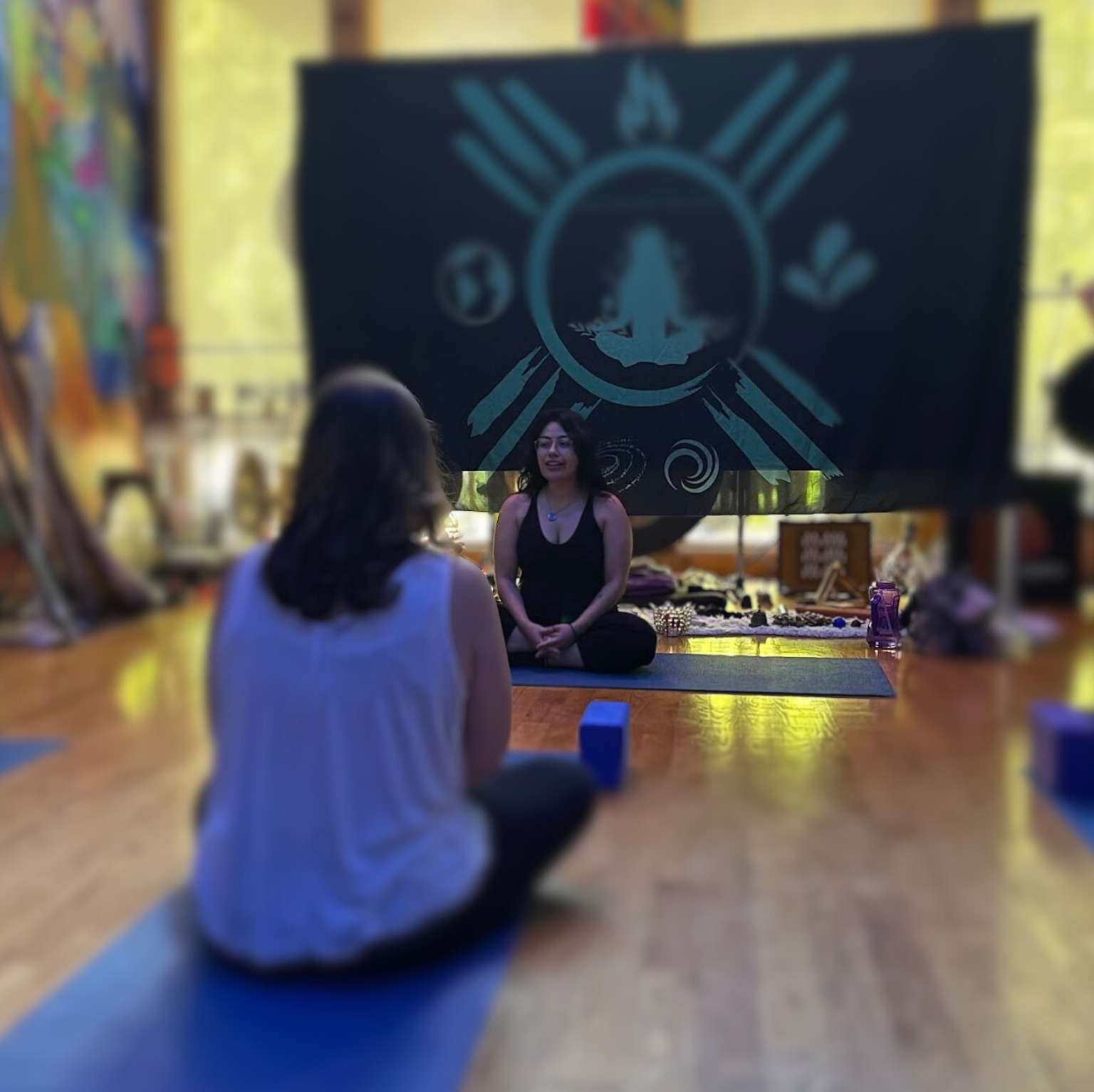 Two women participating in a yoga class in a studio. One woman is sitting cross-legged on a yoga mat facing a large black banner with a meditative figure and symbols on it. The scene is indoors with wooden floors, and various items are visible in the background.