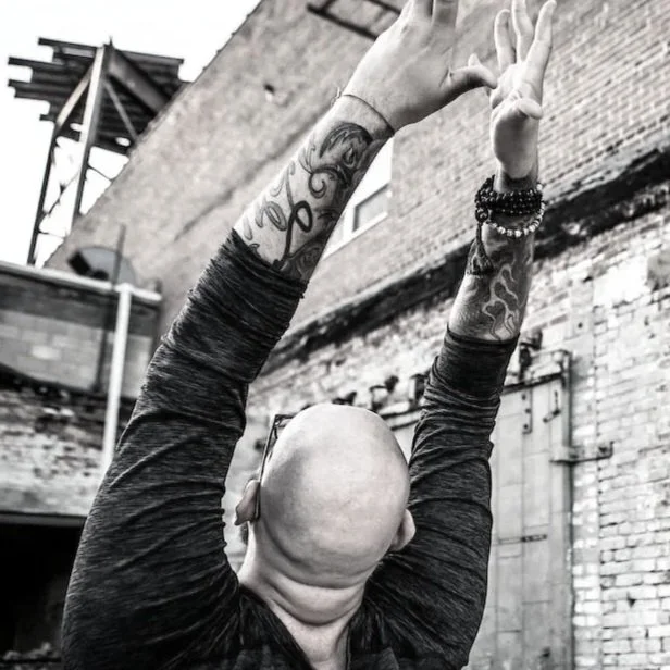 A bald person with tattoos on their arms raising their arms in front of an industrial brick building.