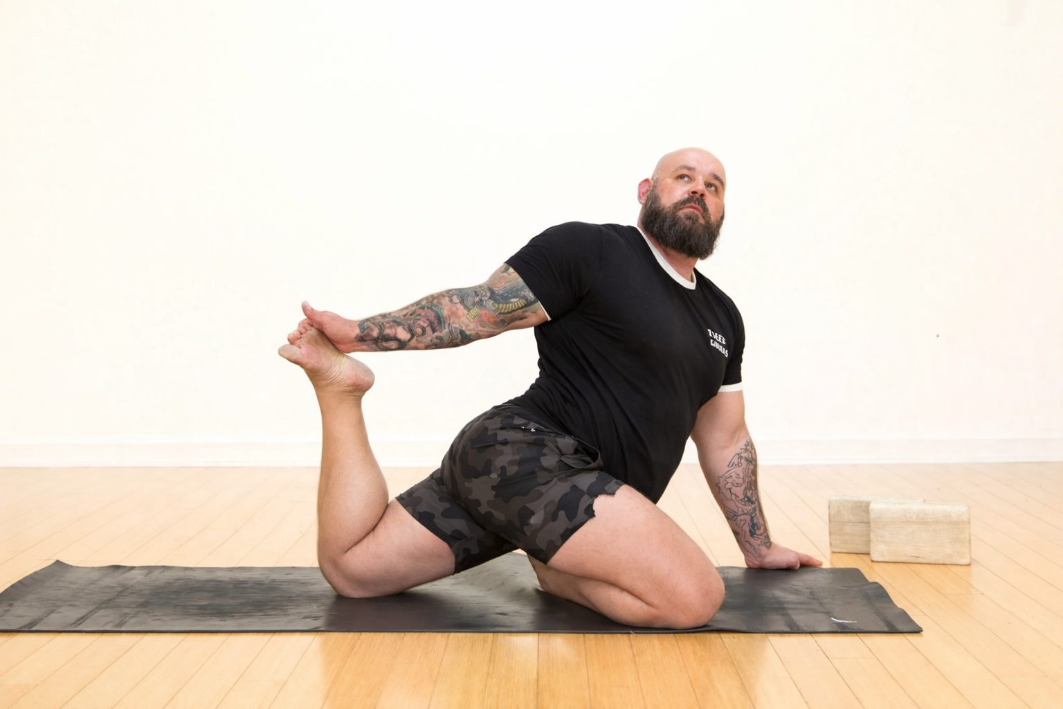 A man with tattoos, wearing a black t-shirt and camouflage shorts, practicing yoga on a black mat in a room with wooden floors and plain white walls. He is in a modified lunging stretch, holding his left foot with his left hand while supporting himself with his right hand on the ground.